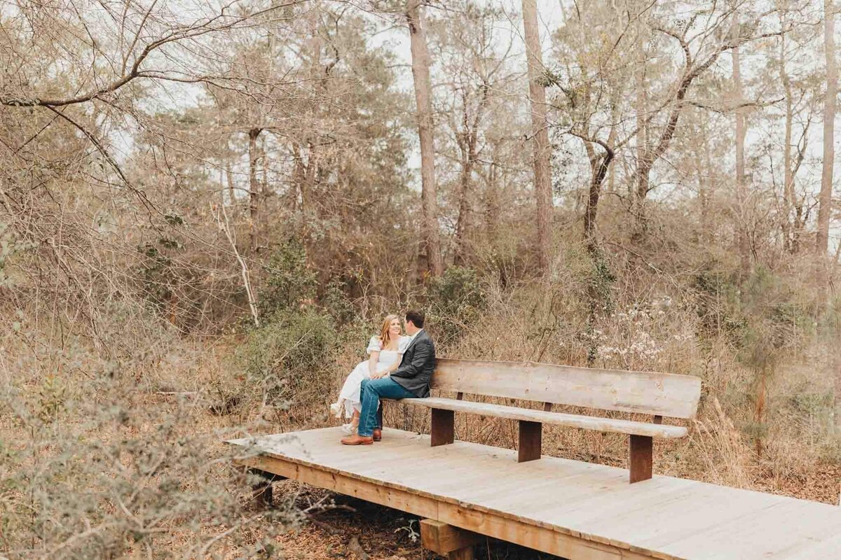 Houston engagement session where couple sits on a long wooden bench in the middle of the woods, talking to each other.