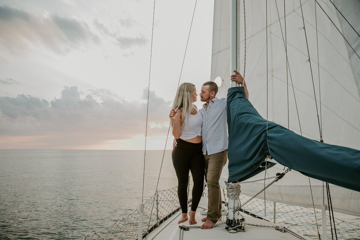 Surprise engagement and photo shoot in Sarnia, Ontario on a sailboat. The man and woman are standing at the bow of the boat embracing. the man has one arm casually wrapped around the mast.