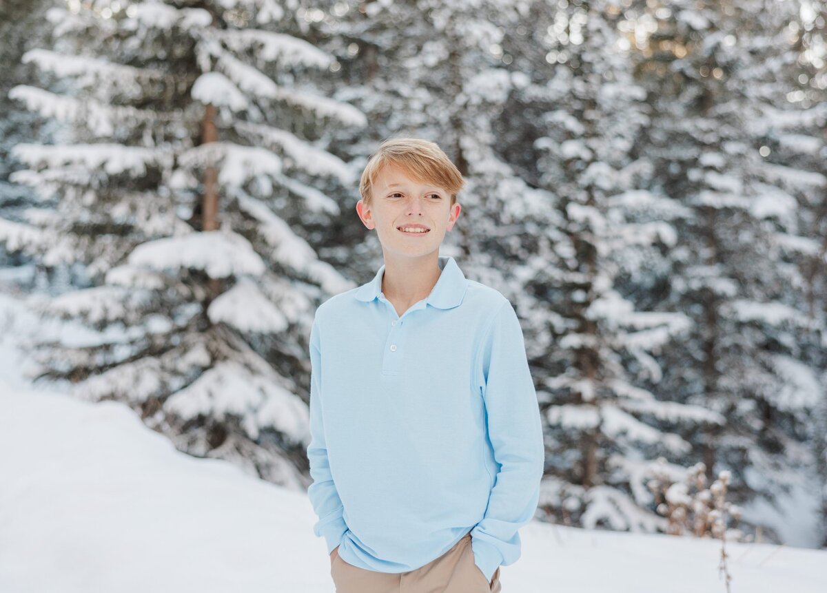 A young teen boy in a blue shirt smiles while standing in a snowy forest 