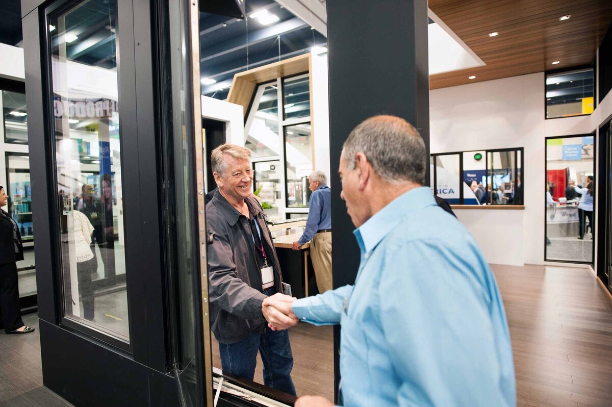 Two men shake hands through a large door at a homebuilders conference