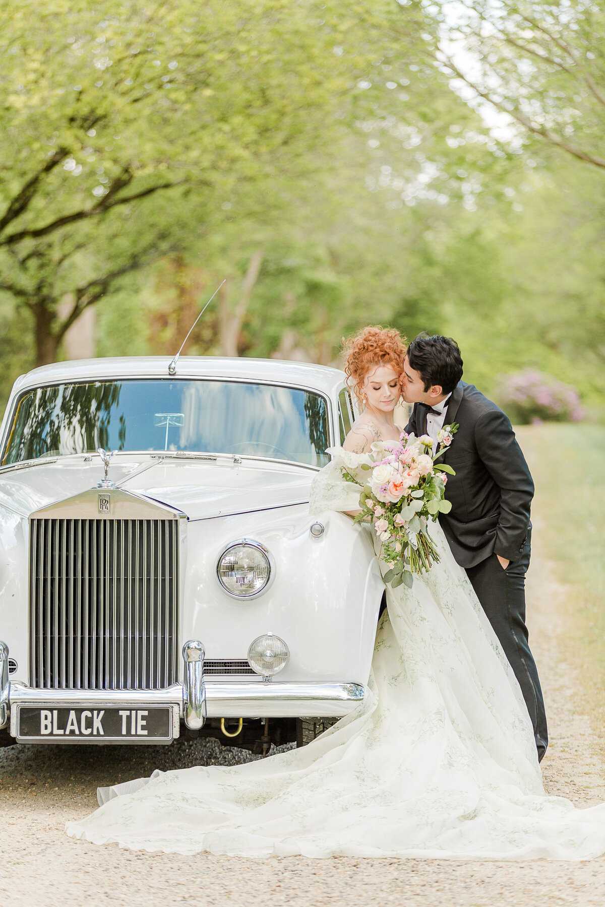 A bride leans against a vintage Rolls Royce at an estate in Boston's North Shore. The groom is leaning in and kissing the bride on her cheek. Captured by best Massachusetts wedding photographer Lia Rose Weddings