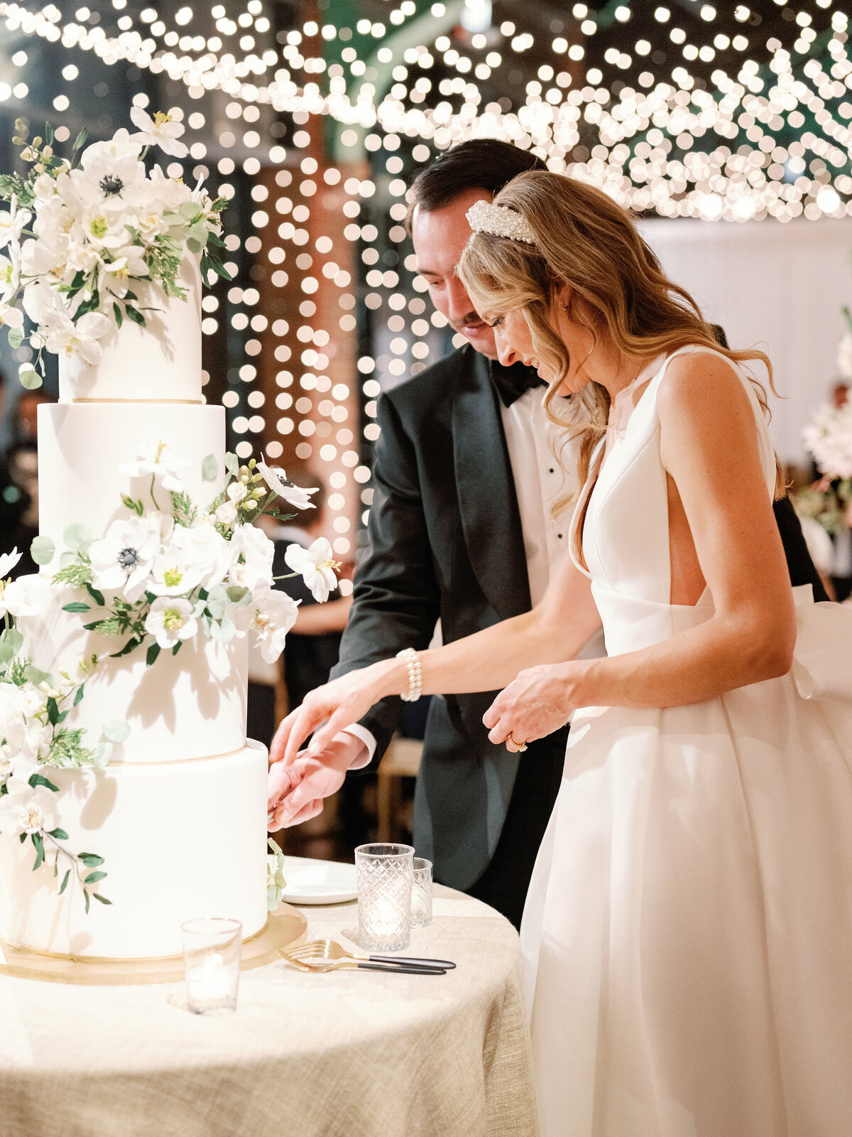 A newlywed couple elegantly cuts their tiered white wedding cake, surrounded by twinkling lights and floral décor.