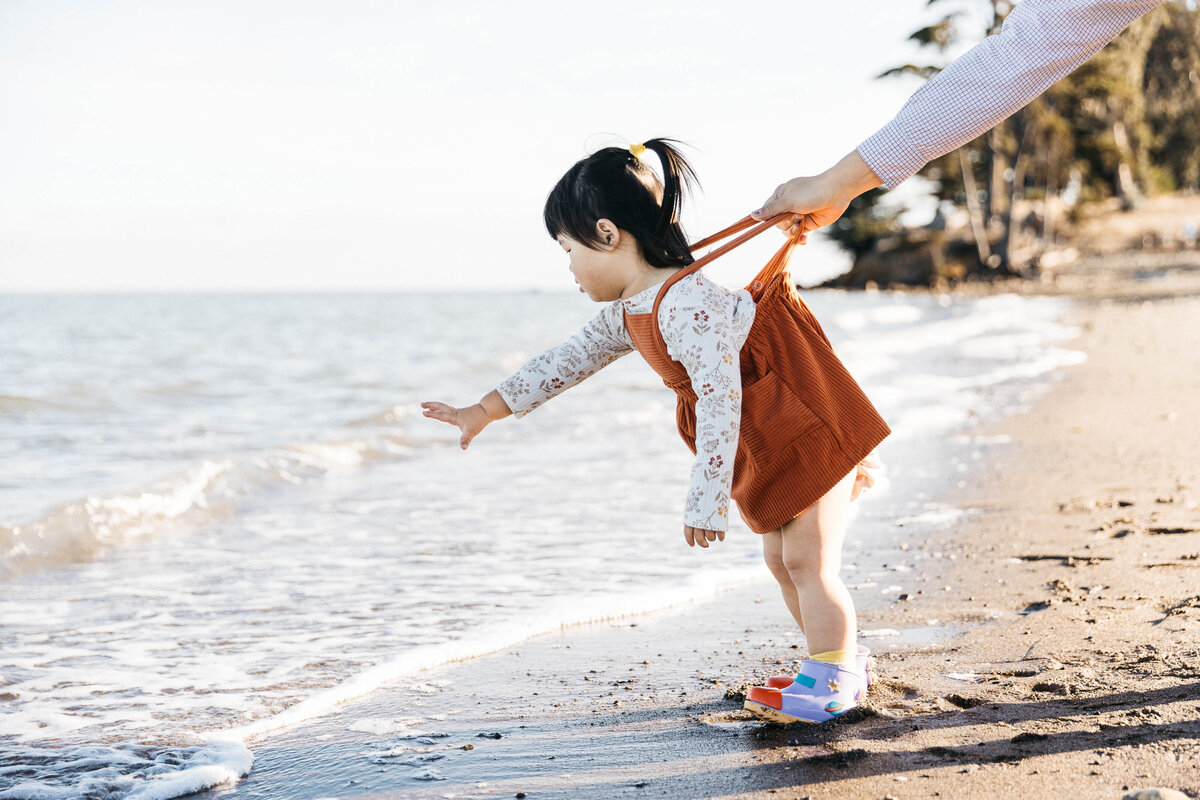 Dad is holding his toddler's suspenders while she leans into the water at the beach.