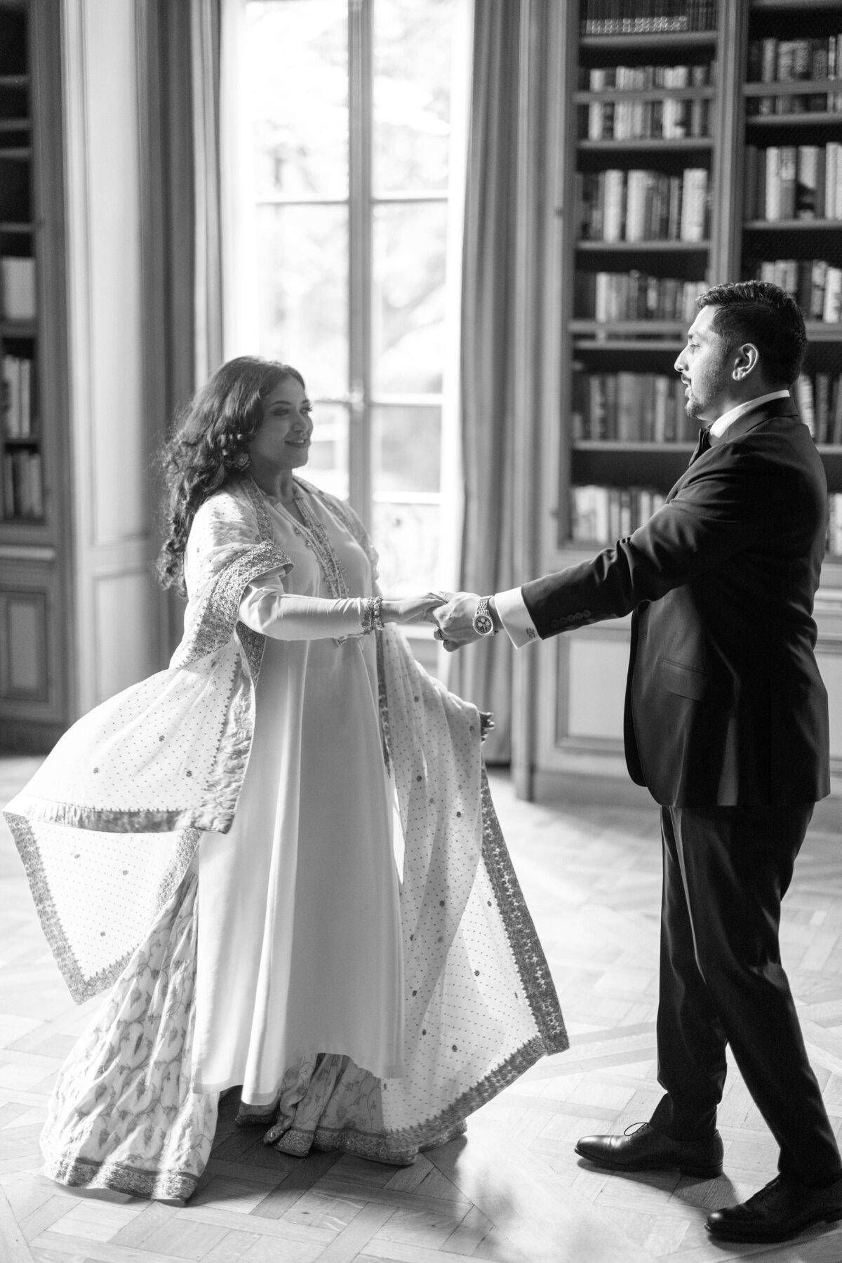 A couple dances in a library, with the woman wearing a flowing saree and the man in a suit. Bookshelves line the walls, and light filters through a window, creating a warm and elegant atmosphere.