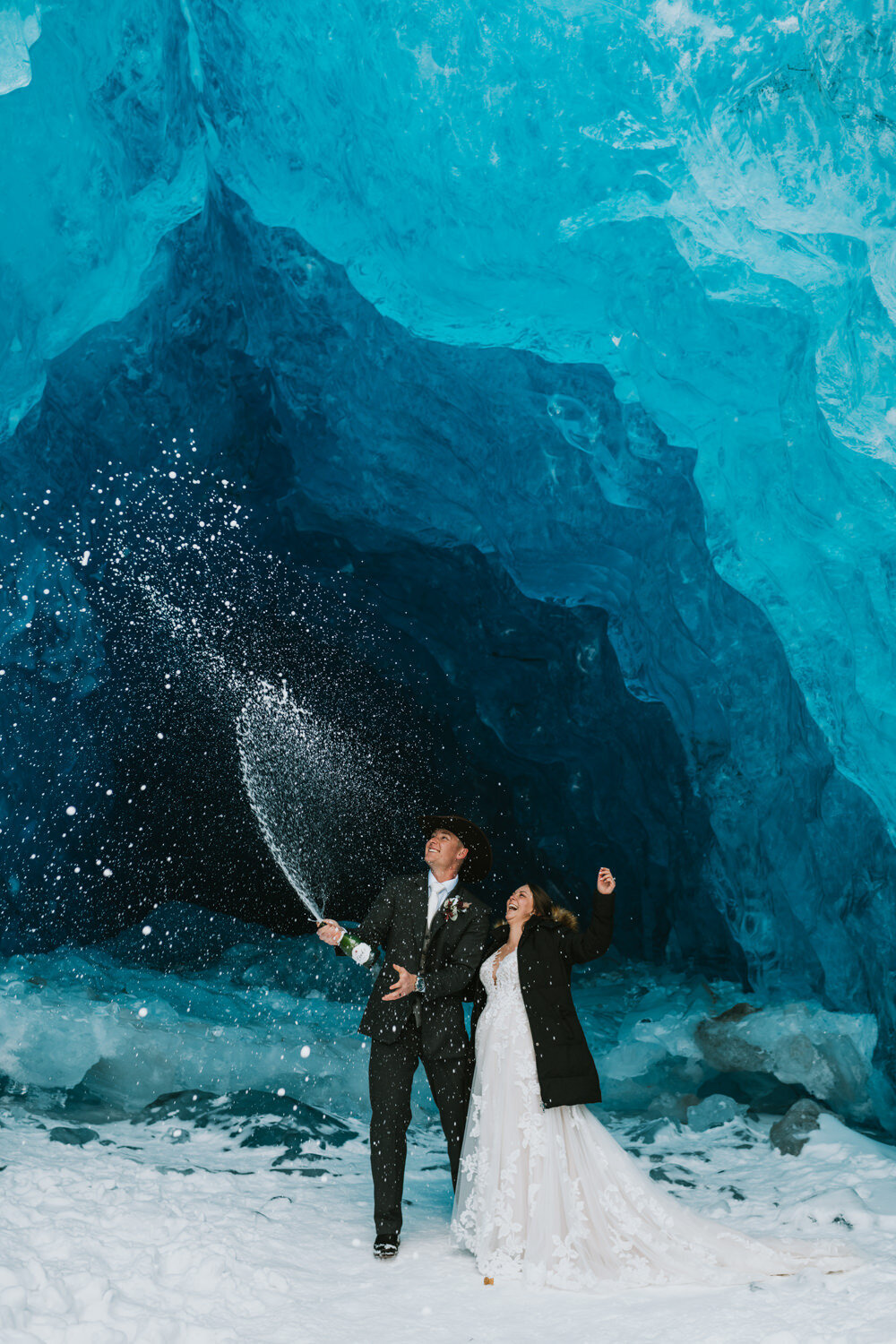 couple in wedding attire standing in a blue glacier ice cave spraying a bottle of champagne on their alaska helicopter elopement