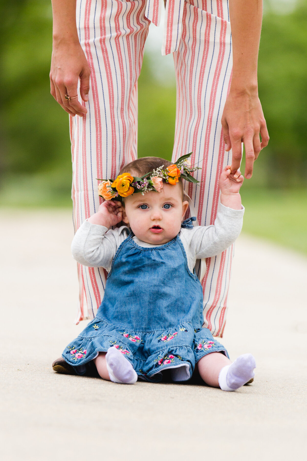 a baby girl sits in front of her mom's legs while wearing a floral crown