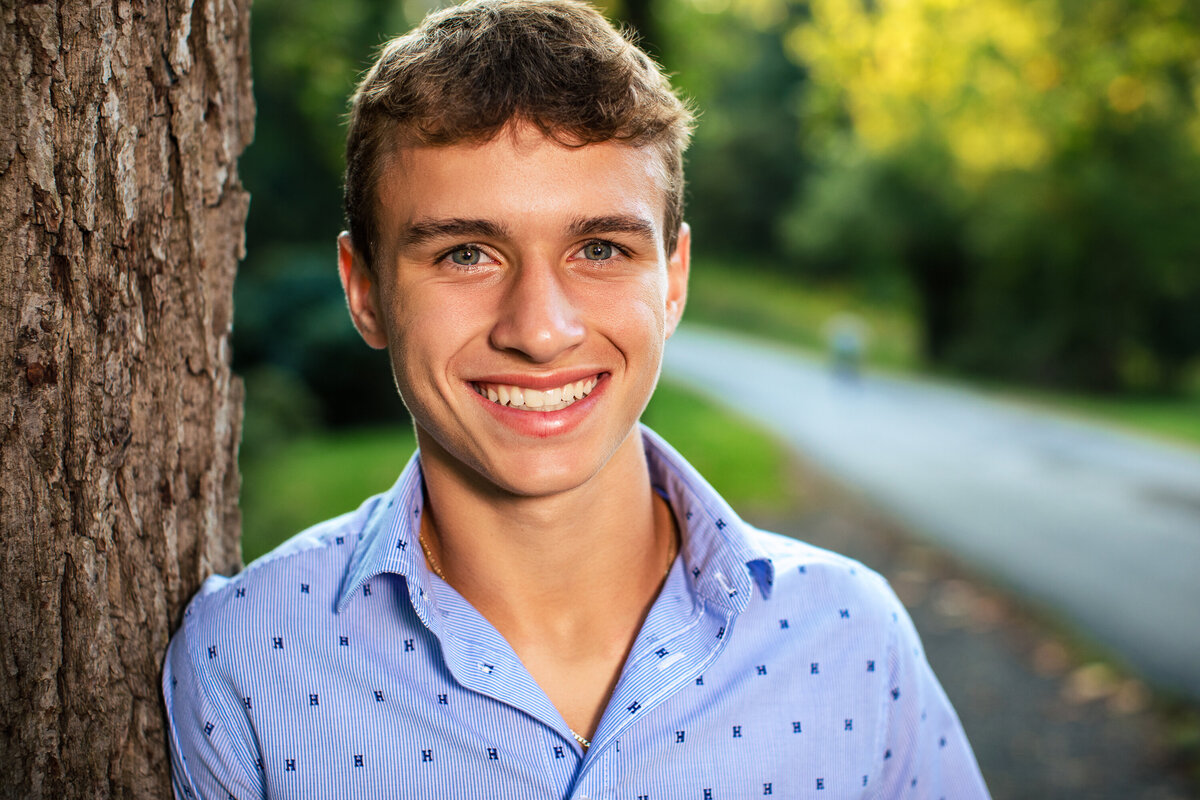 This blue eyed brown haired boy leans on a tree in a park near Pittsburgh Pennsylvania for his senior portraits