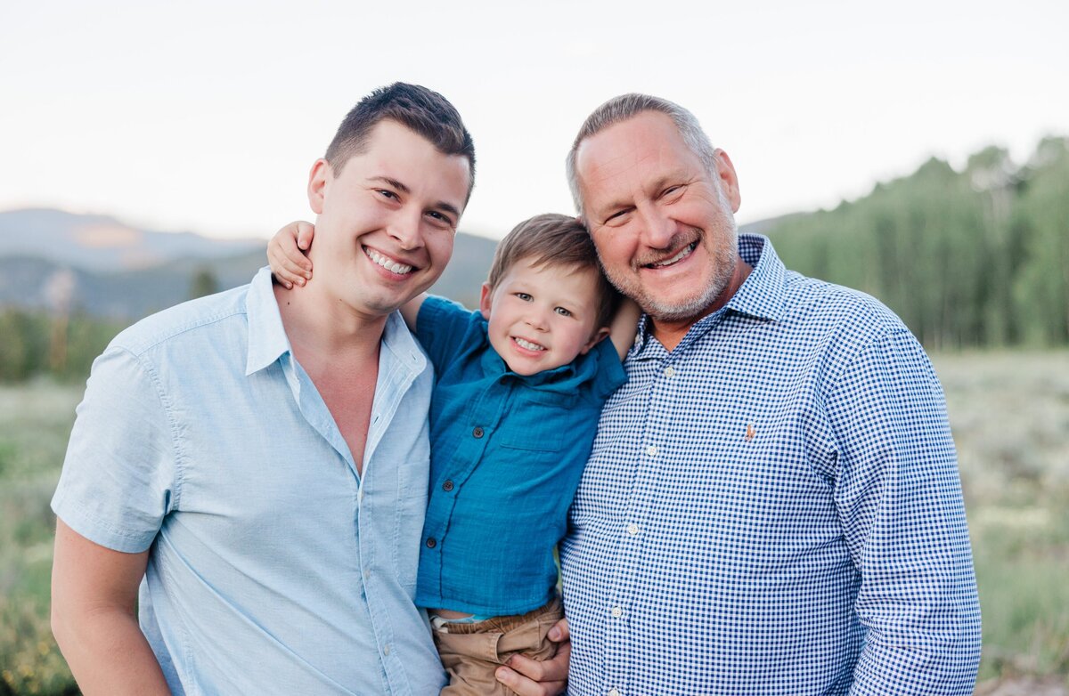 A toddler boy hangs on dad and grandpa while smiling in the mountains in blue shirts