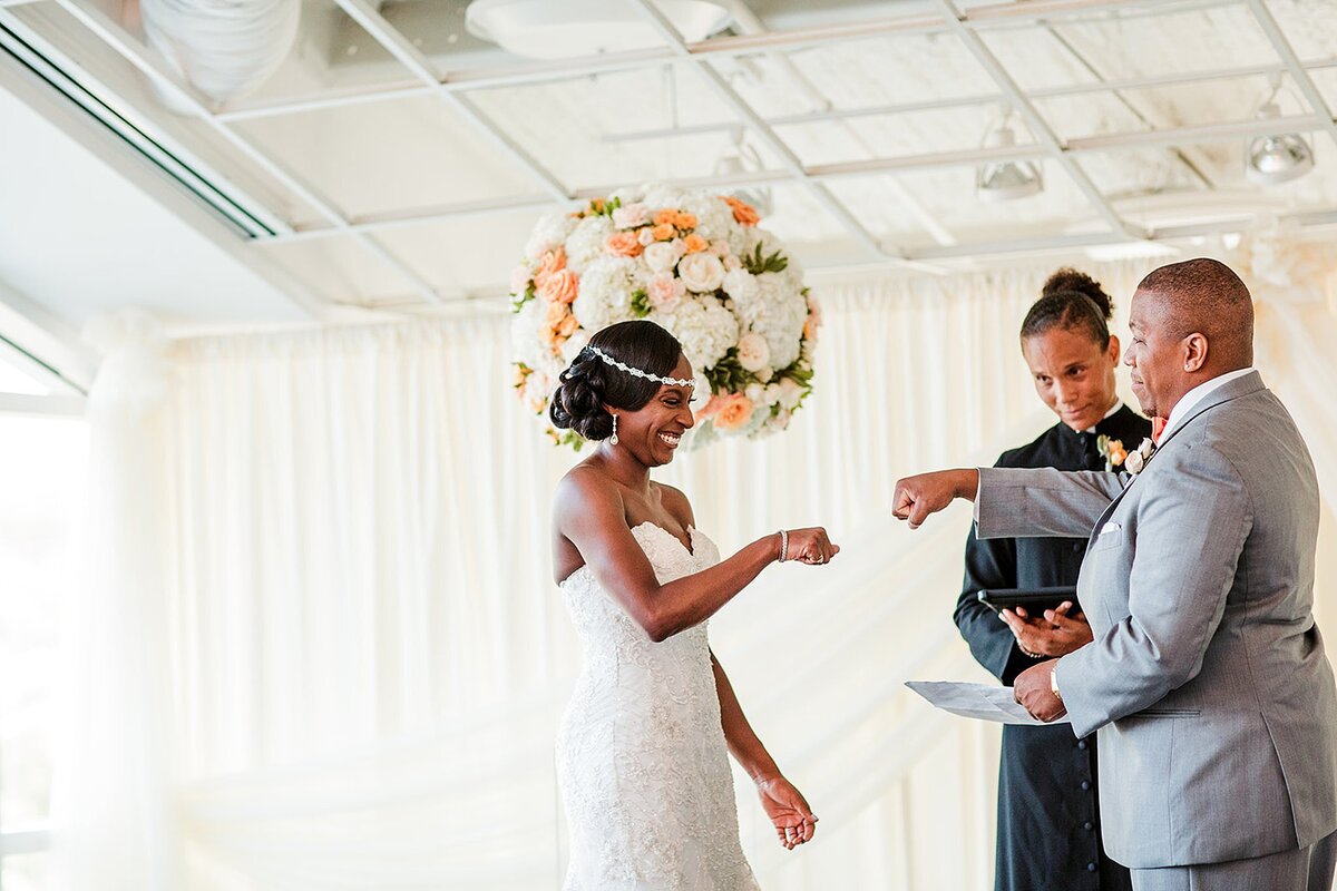 African American bride wearing a strapless wedding gown and a rhinestone tiara fist bumps the groom as he reads his vows. The African American groom, wearing a light gray suit fist bumping the bride  as the African American minister wearing black robes looks on. They are standing in front of sheer white drapery and a large round floral arrangement made of white and ivory flowers accent with peach and orange flowers at The Liff Center Nashville Opera