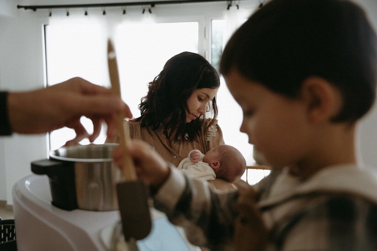 La mère regarde tendrement son bébé dans ses bras, tandis que son fils cuisine avec son père au premier plan. Séance naissance par Kamescop, photographe en Bretagne.