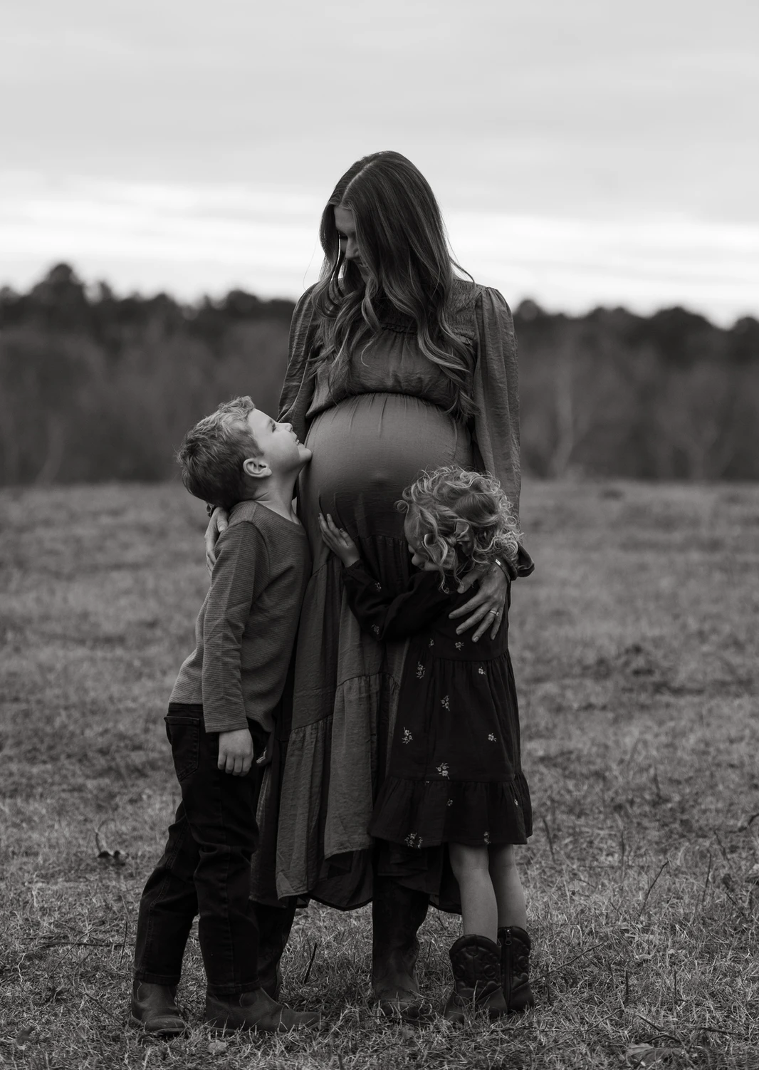 Black and white maternity portrait—mother standing in a field with her children embracing her belly in a quiet, emotional moment.