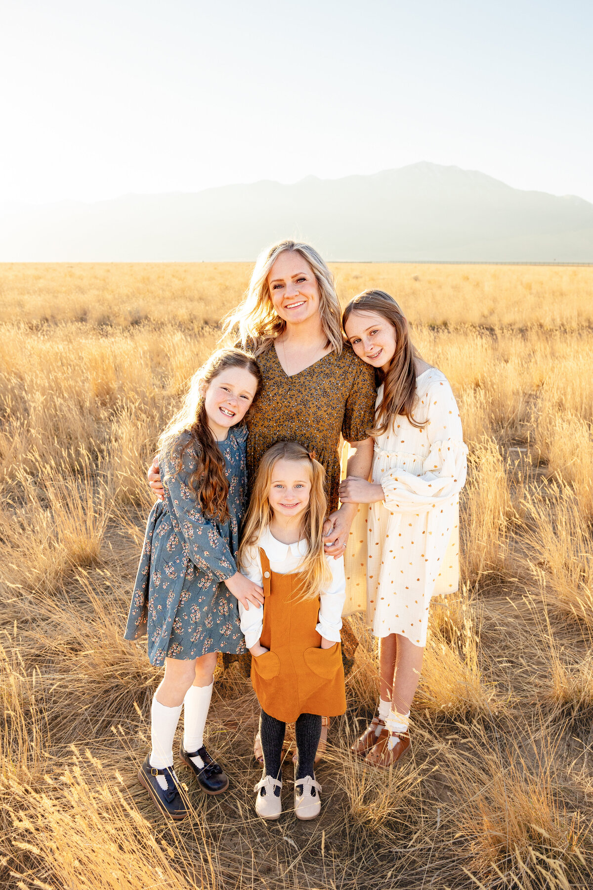A mother stands with her three daughters, all smiling warmly as they pose in a golden field during sunset
