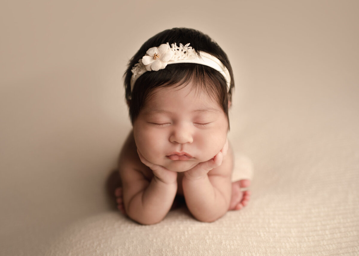 Baby girl in a froggy pose for her Murrieta newborn photoshoot. Baby girl is folded forward with her hands resting under her chin. She is positioned atop of a light beige stretch fabric and wearing a matching headband. Baby's chin is resting atop of her hands, propping her up. Captured by best Murrieta newborn photographer Bonny Lynn Photography