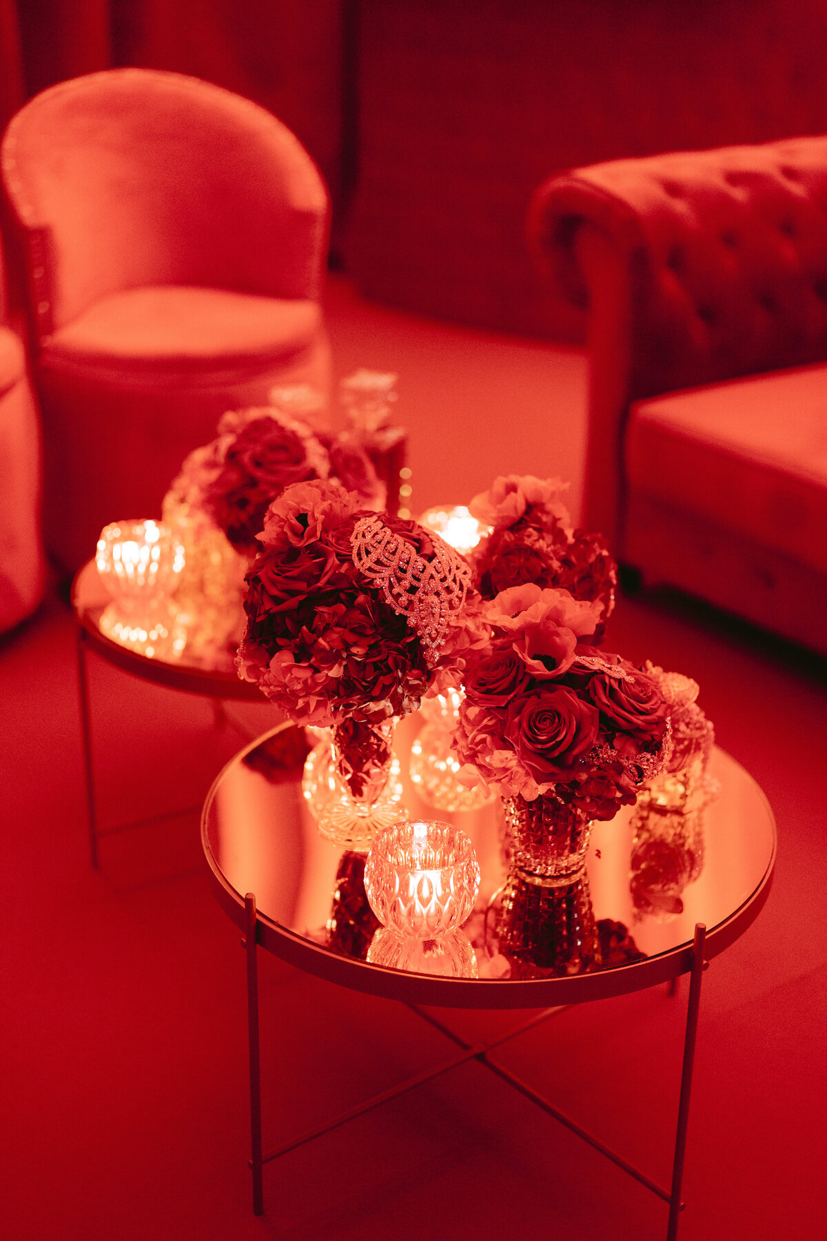 A cozy, red-lit room with an elegant setup. The image features two plush chairs, a tufted sofa, and a glass table adorned with bouquets of red and pink flowers and small, glowing candles. The ambiance is intimate and warm.