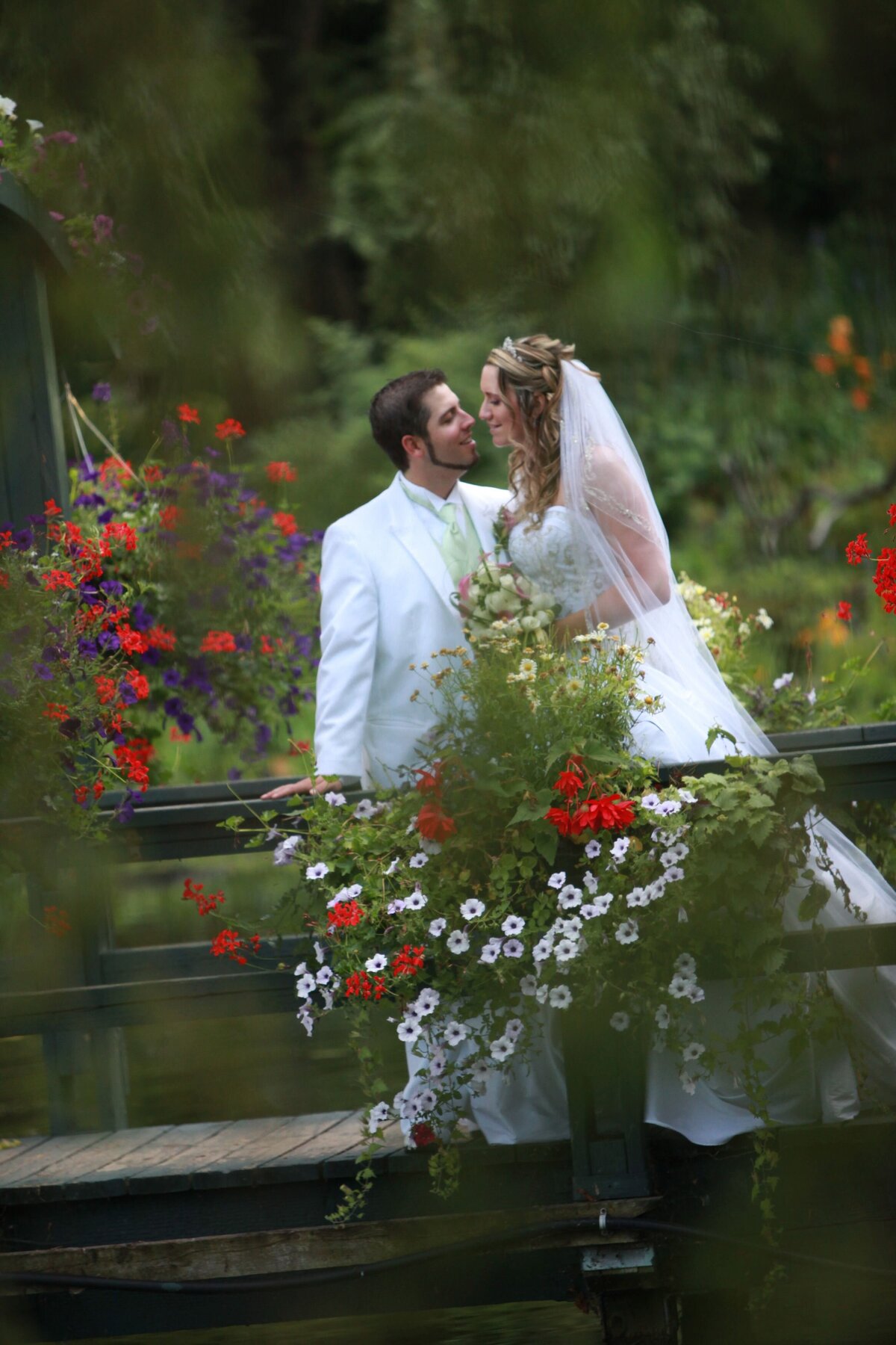 The bride and groom pose together beside vibrant red flowers. The rich colors of the flowers complement their elegant wedding attire, creating a striking and romantic scene. The couple's joyful expressions and close embrace highlight their love and the beauty of their special day.