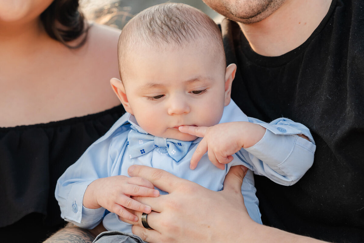 A baby wearing a blue shirt and bow tie sits in his parents lap during a beach photoshoot.