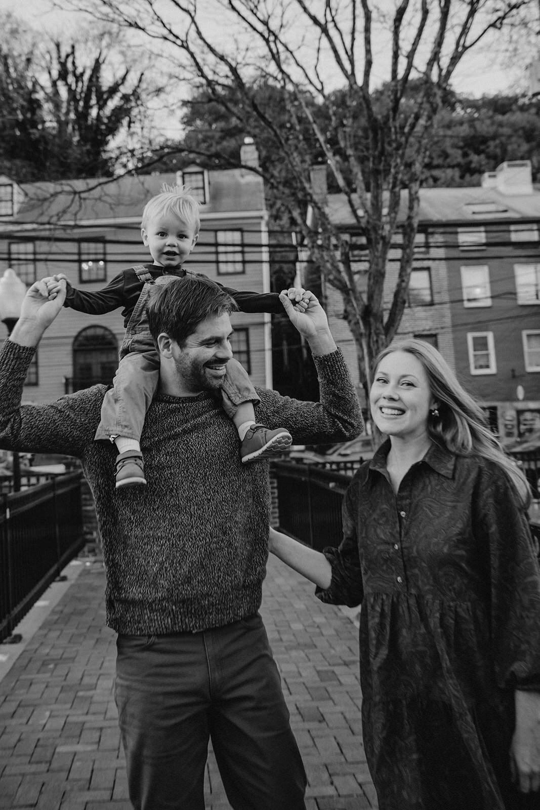 Parents walking along a sidewalk as one holds their child on their shoulders 