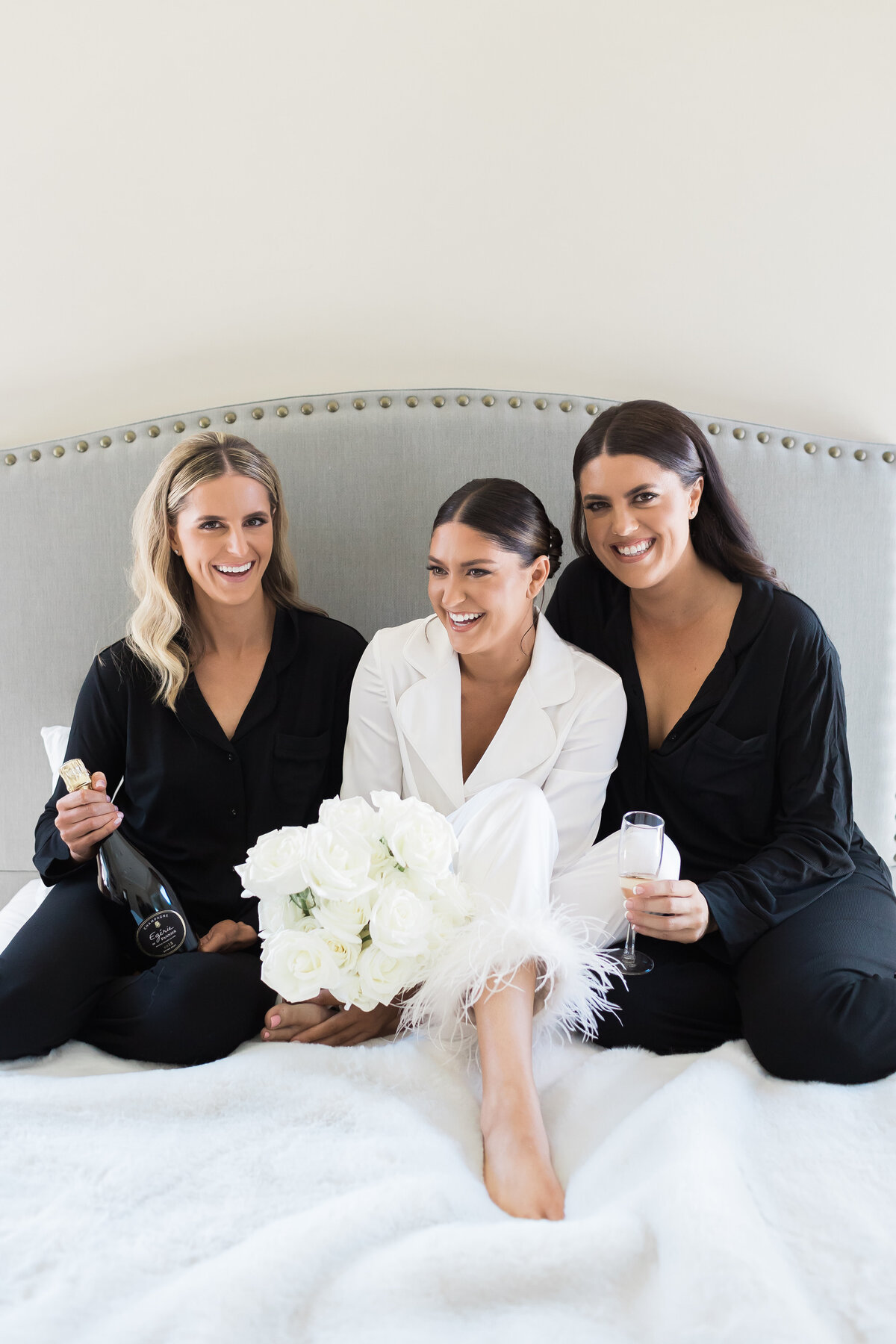 Chic bride laughs on a bed drinking champagne with her bridesmaids