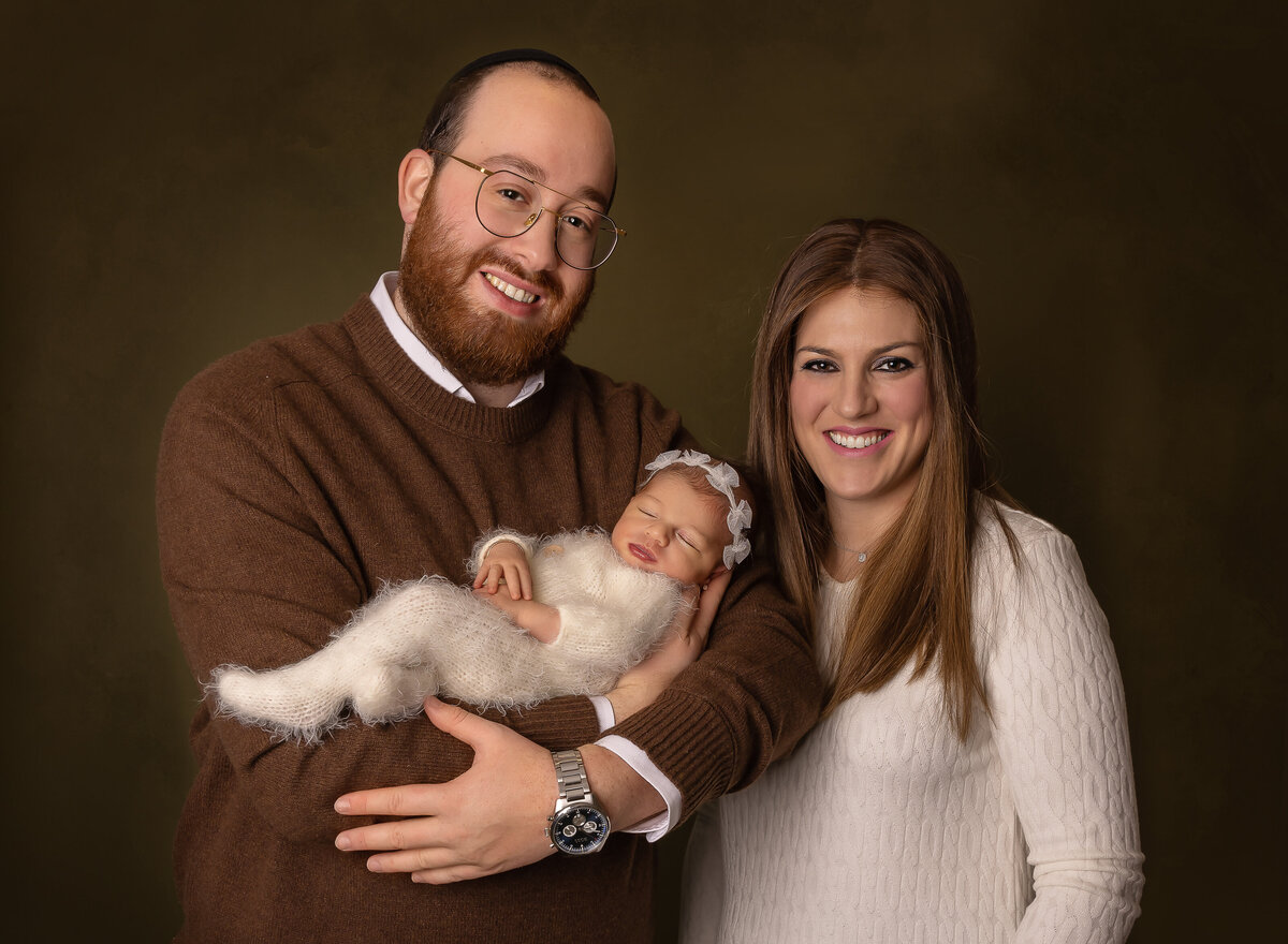 Mom and dad pose with baby for brooklyn newborn photoshoot. Dad is holding baby, baby is wearing a white onesie and headband. Mom and dad are smiling at the camera as baby sleeps. Captured by top Brooklyn newborn photographer Chaya Bornstein