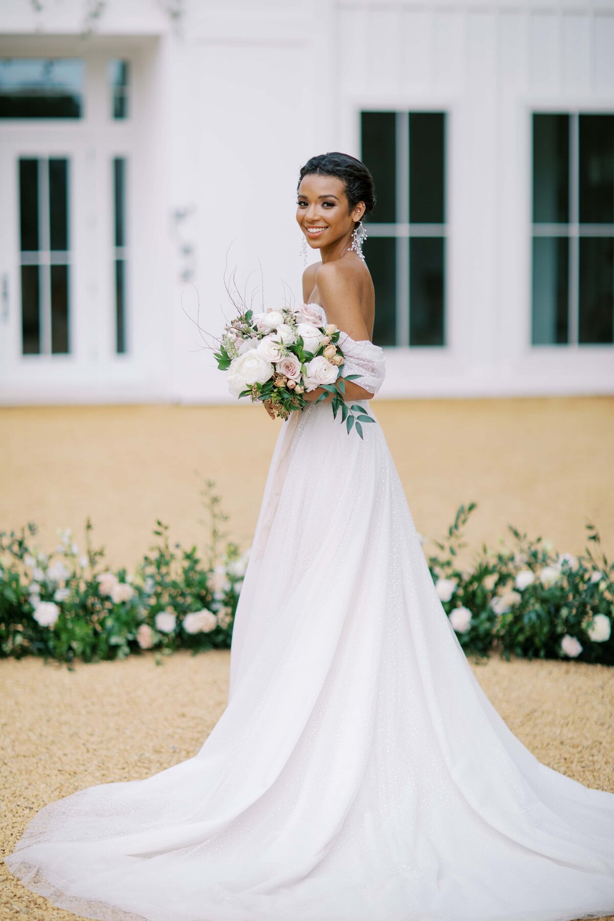Smiling black bride in a gorgeous wedding gown holding a bouquet of white and pink flowers in front of the Ivy Rose Barn