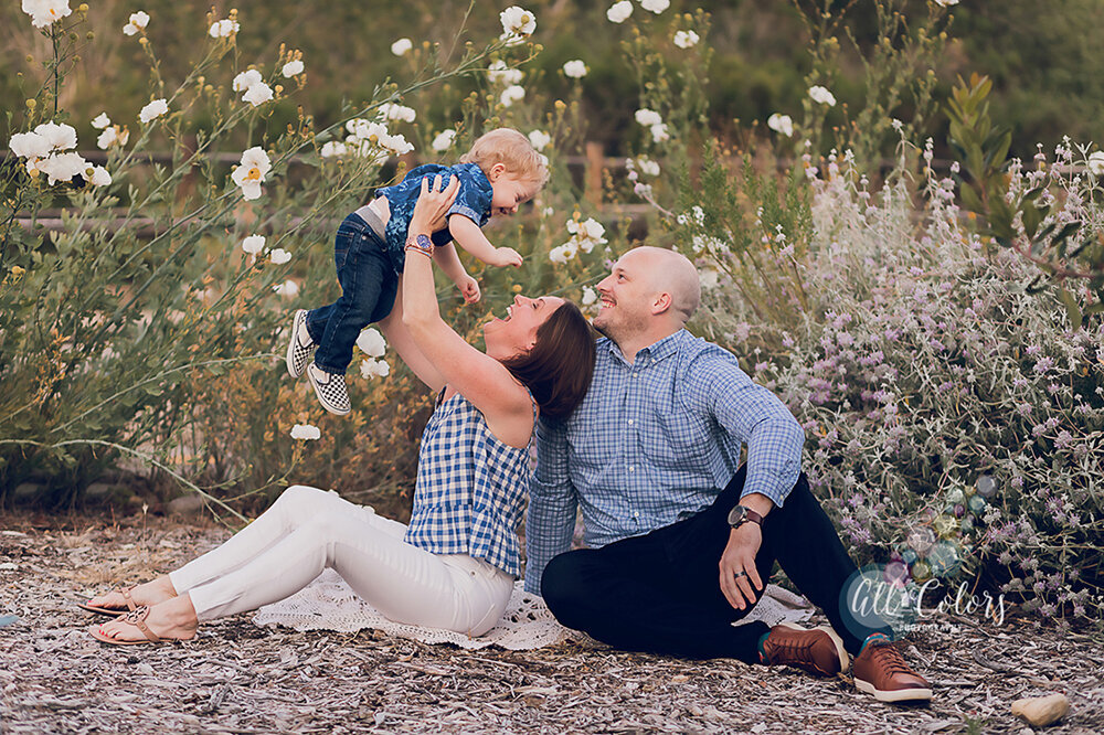 photo of a mom, dad, and toddler. They are seated and mom is lifting the child up over her head. White poppy flowers and sage in the background