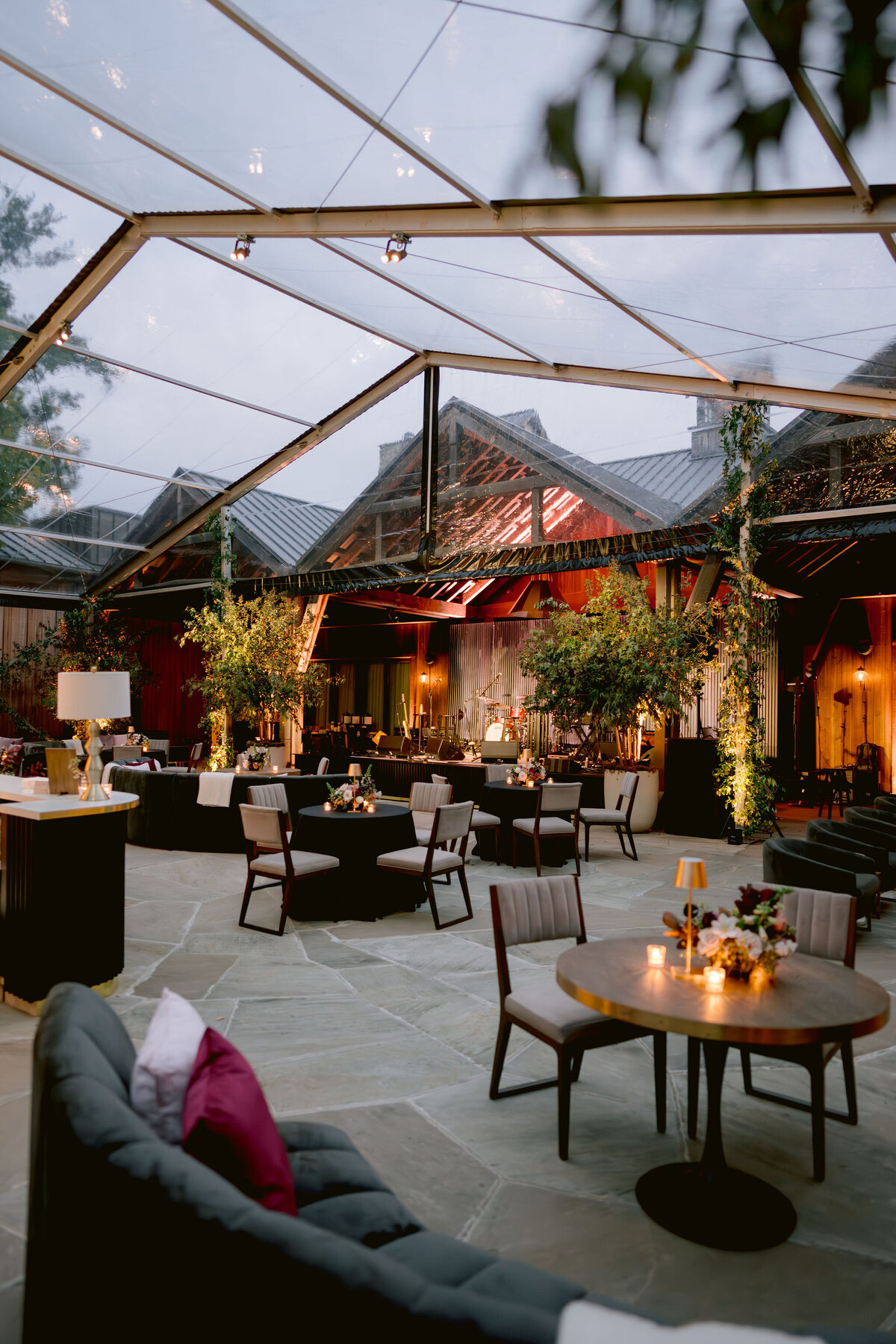 Moody welcome party for fall wedding weekend. Tented reception for outdoor wedding reception at Blackberry Farm. Moody floral design with colors of deep magenta, mauve, eggplant, blush, and natural greens. Rich tones with roses, mums, and smokebush foliage. Romantic candle light and large trees create a magical setting for a welcome party. Destination wedding at Blackberry Farm. Design by Rosemary & Finch Floral Design in Nashville, TN.