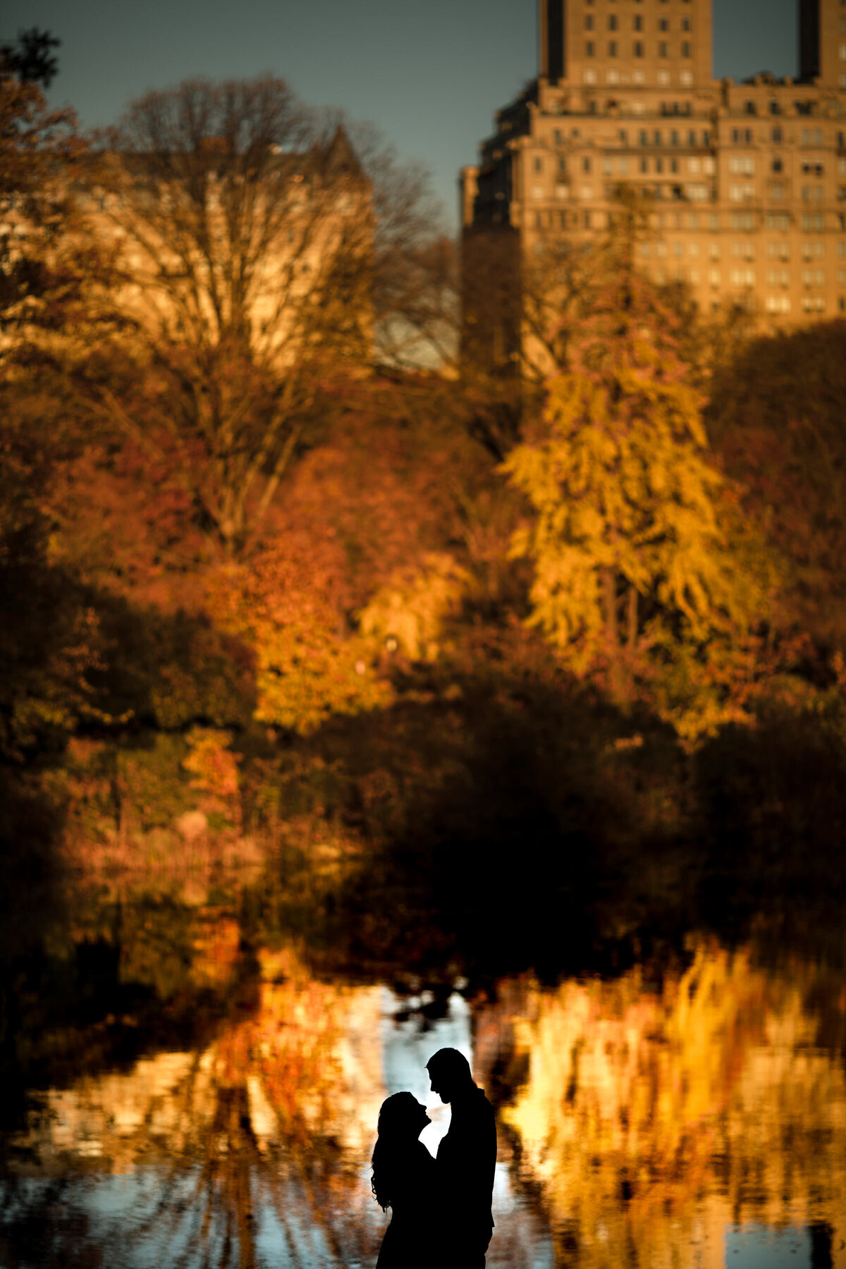 A couple about to kiss in front of a lake surrounded by fall colored trees.
