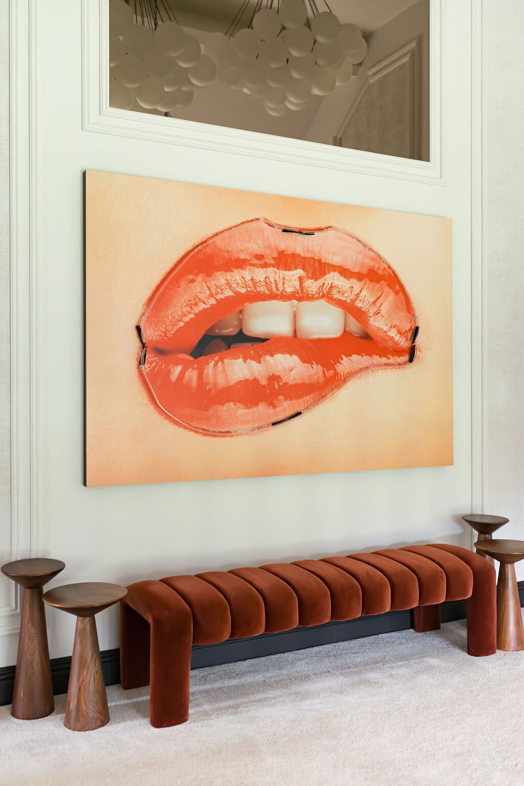 A large, vibrant painting of red lips biting the lower lip is displayed on a white wall. Below the painting is a cushioned, burnt orange velvet bench flanked by two wooden side tables. The room features a modern and minimalistic design with neutral tones.