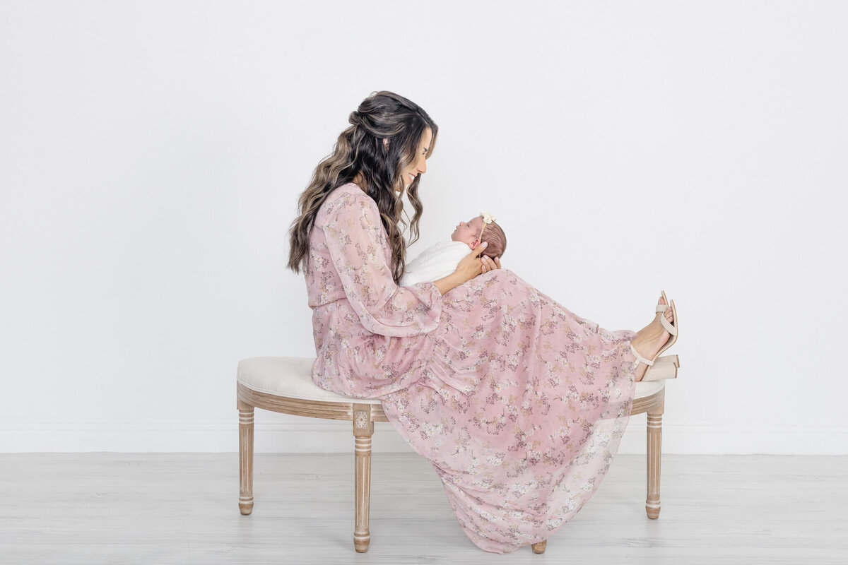 A woman in a flowing pink floral dress sits on a bench, holding a baby wrapped in a white blanket. The scene is set against a plain white background, and the woman gazes lovingly at the baby.