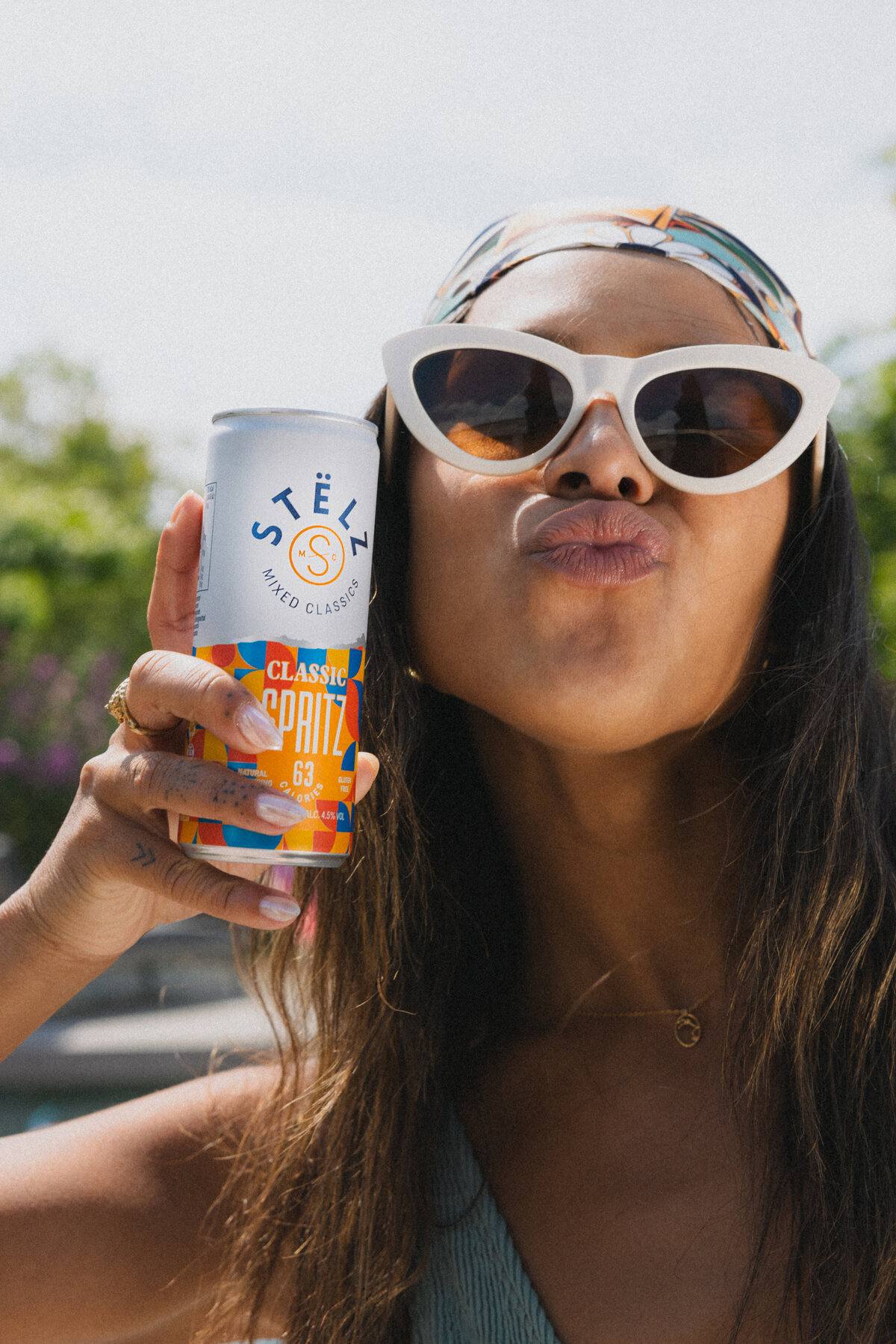 A woman with a playful smile poses for the camera while holding up a can of STËLZ Classic Spritz. She’s wearing large sunglasses and a colorful headband, creating a festive and summery atmosphere. Photo taken by Bo Eppink.