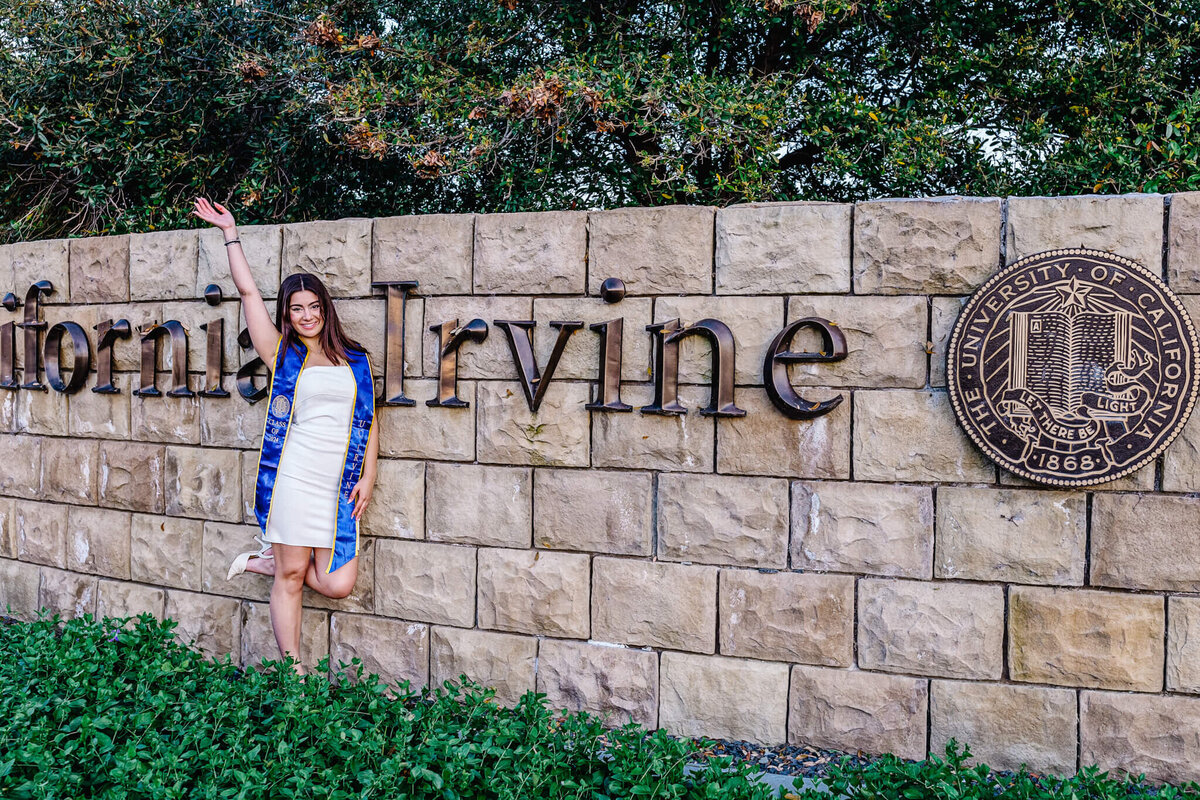 A person in a white dress and blue sash poses with one arm raised in front of a stone wall with the words California Irvine and a circular University of California seal. Green shrubs are in the foreground, and trees are in the background.