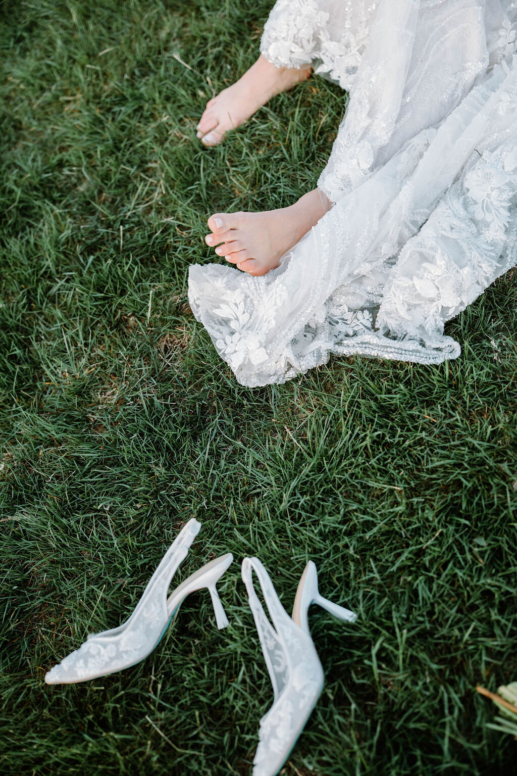 Rhode Island Wedding Photographers | A bride's bare foot peeks from a white lace gown on lush green grass, with clear high-heeled shoes nearby. 