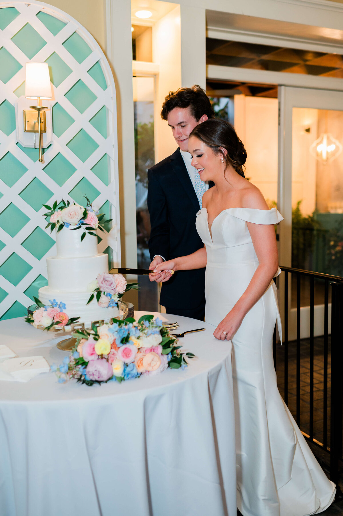 A bride and groom, smiling, cut a white, floral-decorated wedding cake together at a reception. The bride wears an off-shoulder white dress; the groom is in a dark suit. A flower arrangement sits on the table.