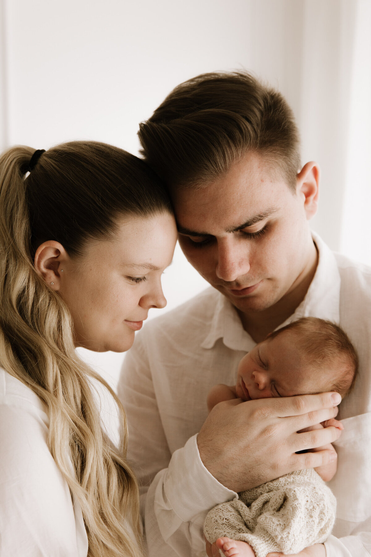 Close-up portrait of new parents gently holding their sleeping newborn in a soft, white studio setting. The father cradles the baby’s head while both parents lean in, foreheads touching, creating an intimate and peaceful moment. The baby is dressed in a knitted cream outfit, and the atmosphere feels calm and full of love.