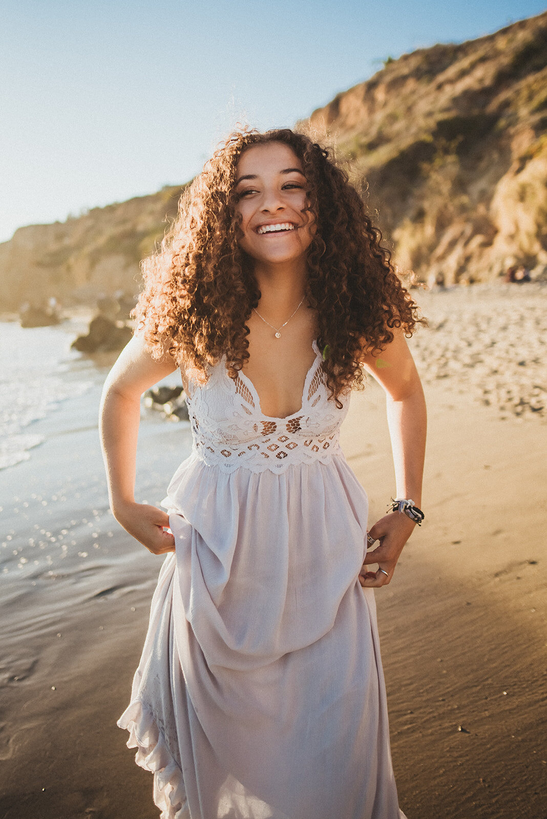 Olivia pulls her white dress out of the water and laughs at El Matador beach.