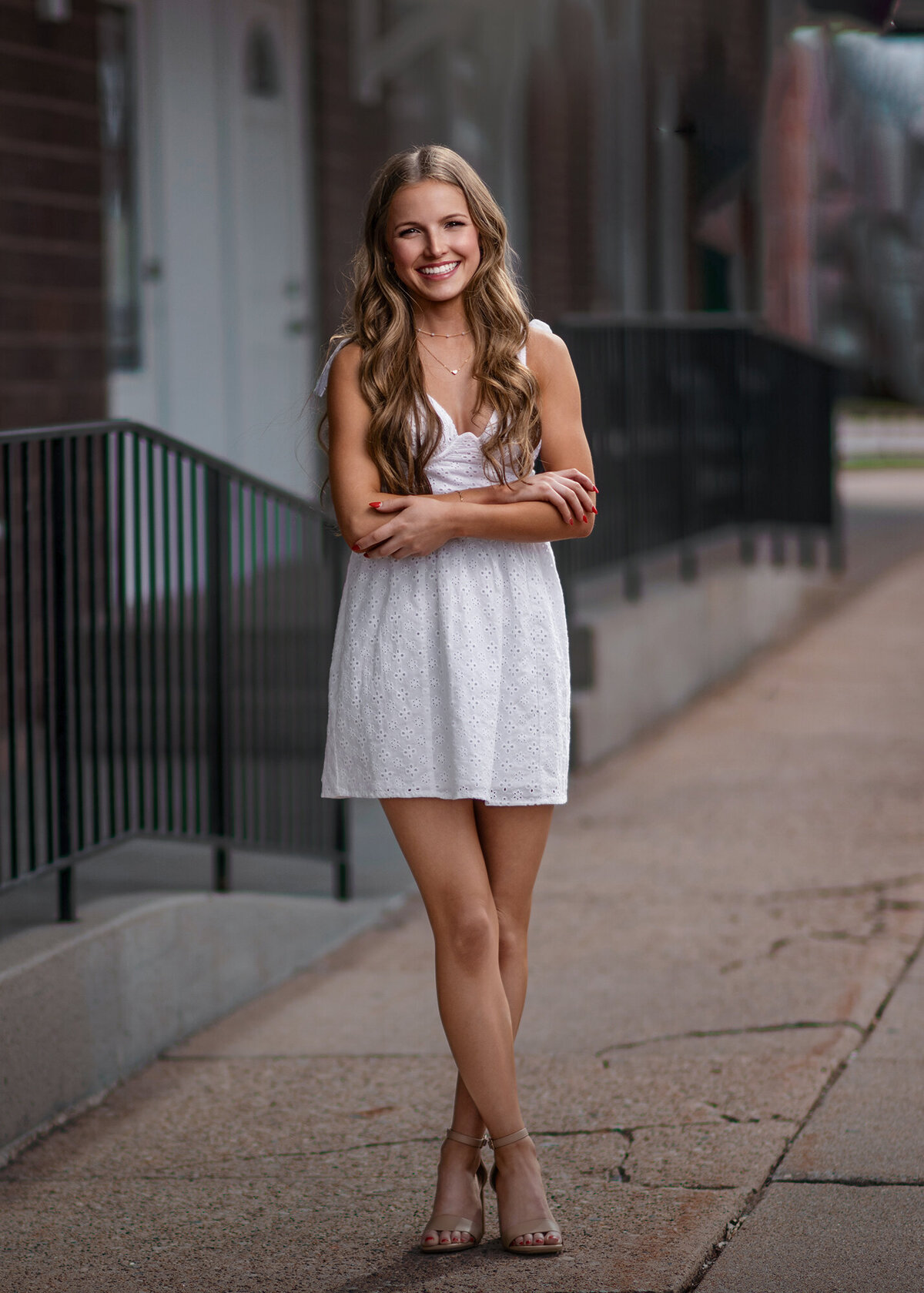 A high school grad standing on a sidewalk in a white dress with arms crossed