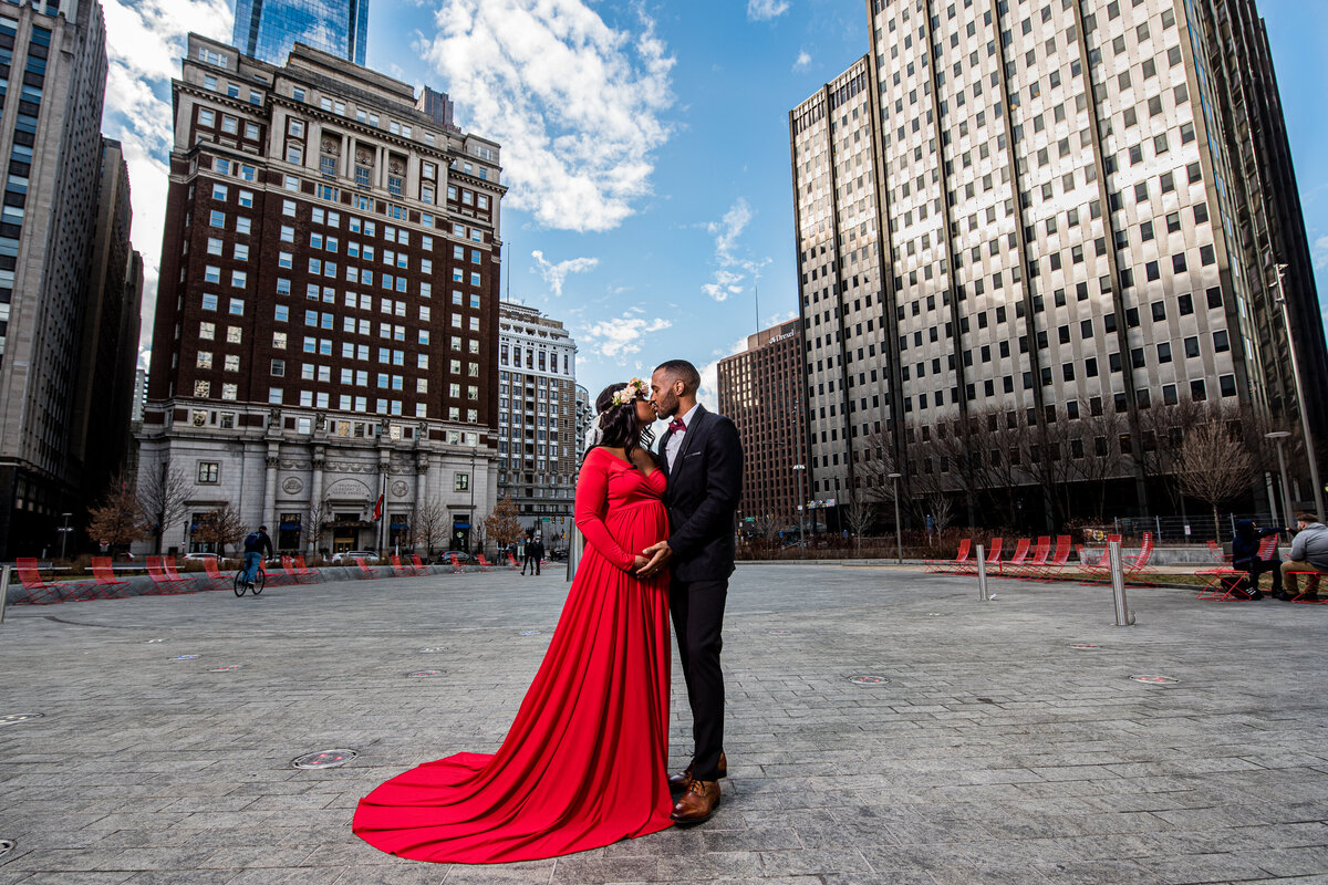 a Black couple kiss in Downtown Austin. She's wearing a red maternity dress and he's wearing a blue suit