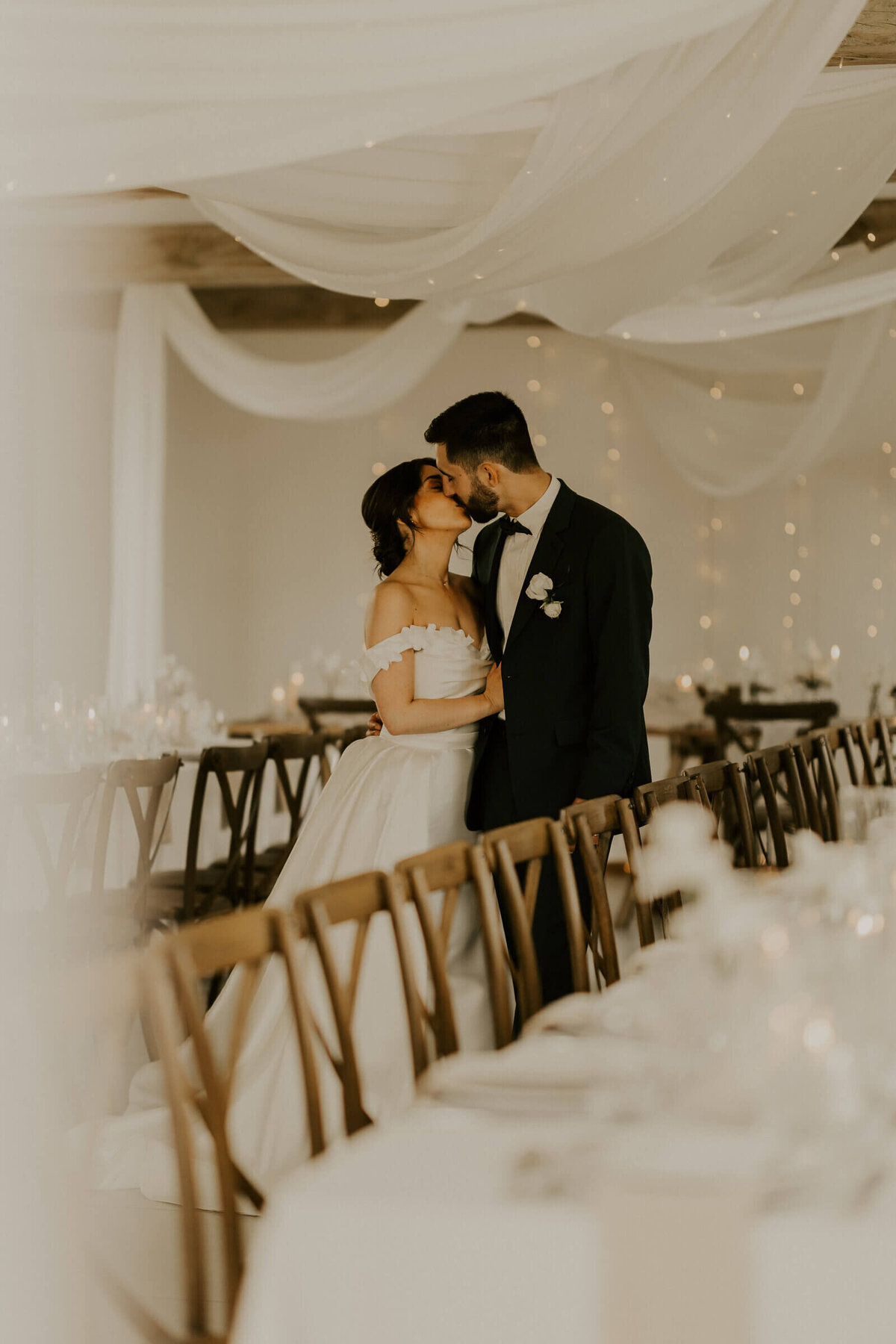 Mariés échangeant un bisou dans la salle de repas vide décorée de voilages, de blanc et de chaises en bois lors d'un shooting photo mariage.