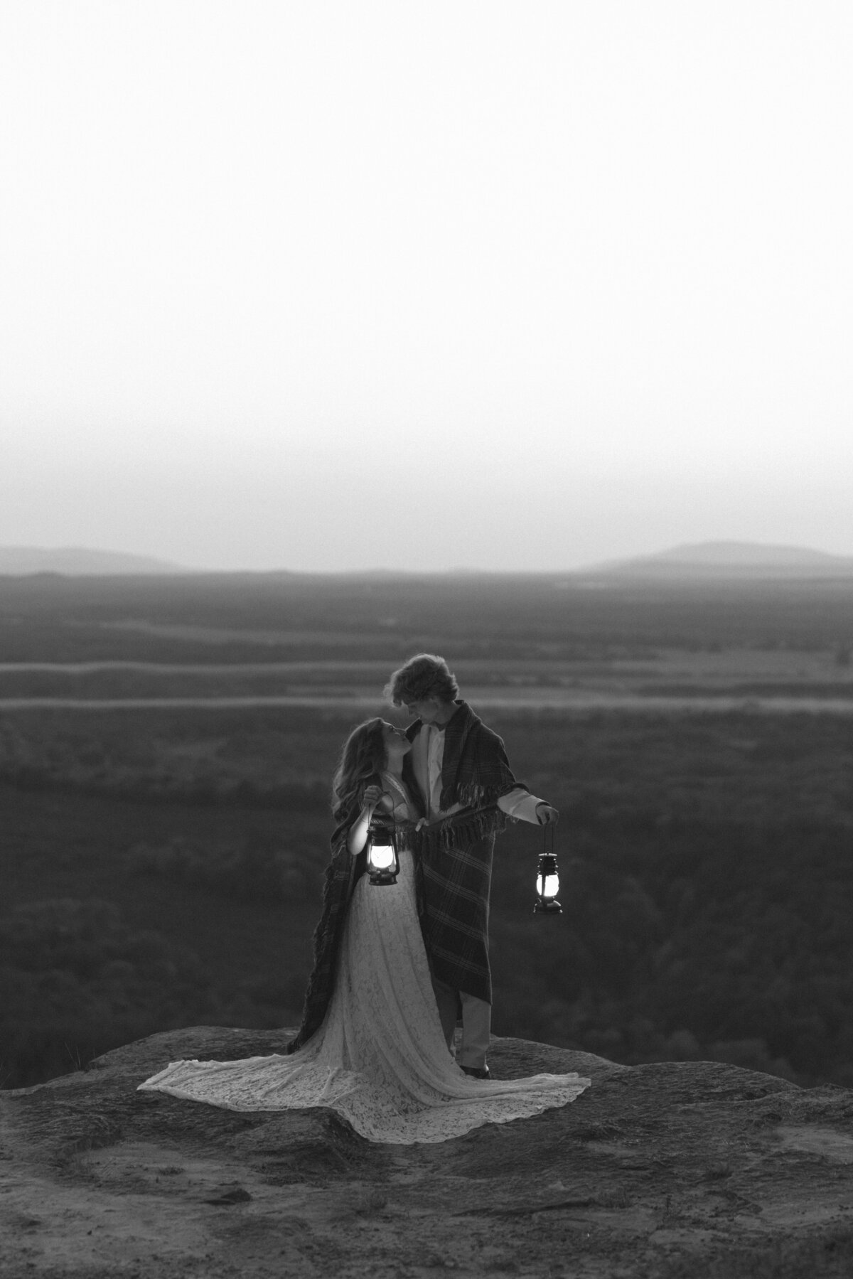 an elopement photo of a couple standing on a cliff at CC Overlook in Petit Jean State Park, Arkansas while holding lanterns during blue hour.