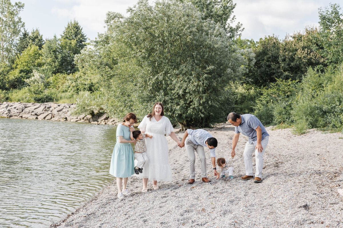 A family gathers on the rocky lakeside shore at Colonel Samuel Smith Park, with trees and bushes framing the scene. Grandparents enjoy moments interacting with small children, creating a perfect snapshot of joy and togetherness.
