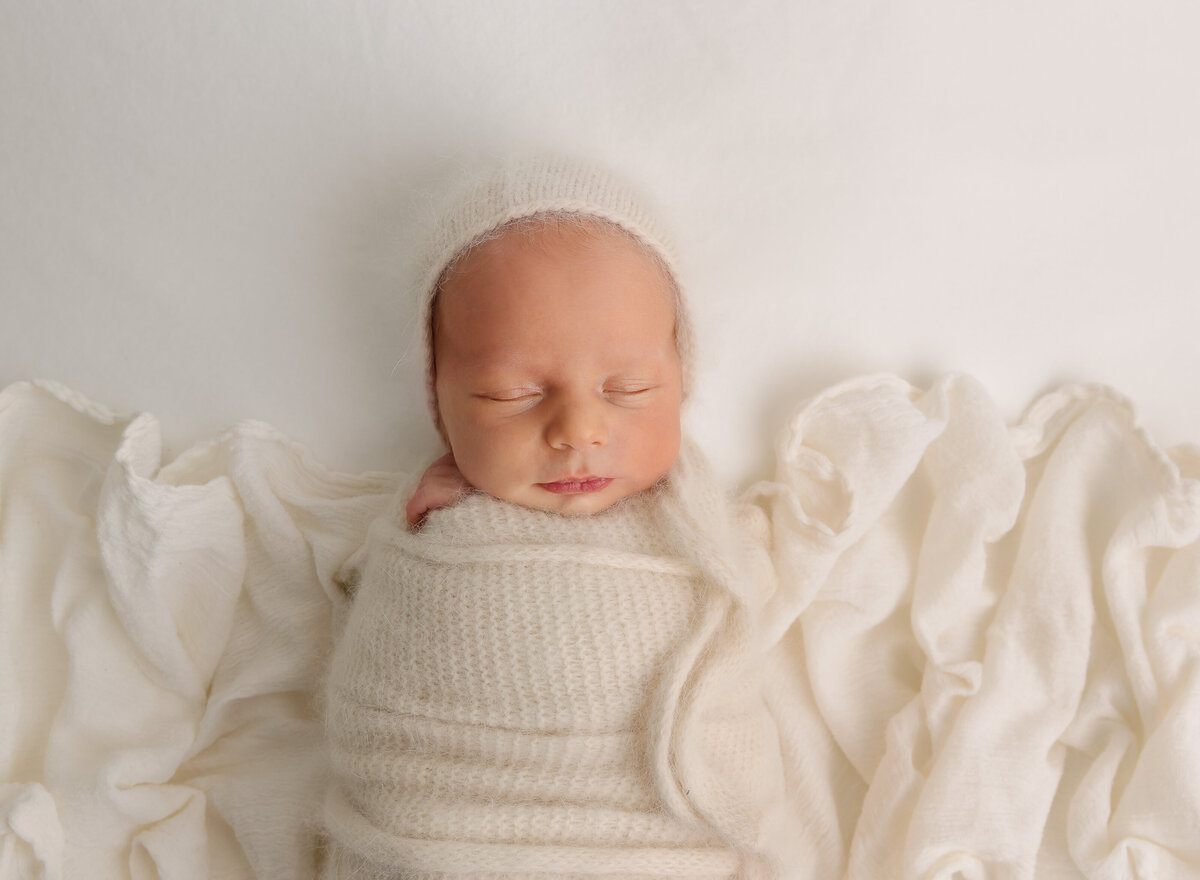 Newborn baby boy is wrapped in cream knit fabric for his brooklyn newborn photoshoot. Aerial image. The baby is placed atop of matching ruched fabric and is wearing a matching knit bonnet. Baby is sleeping. Captured by top Brooklyn newborn photographer Chaya Bornstein