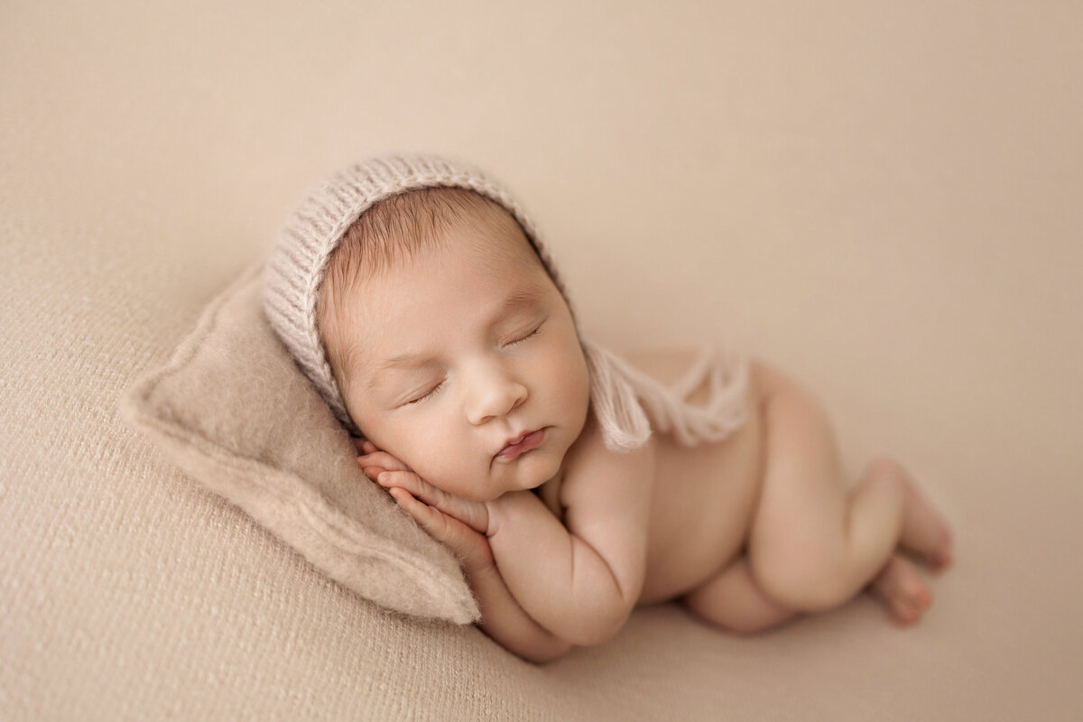 Newborn baby sleeping on a beige pillow, wearing a soft knit bonnet, on a matching beige background.