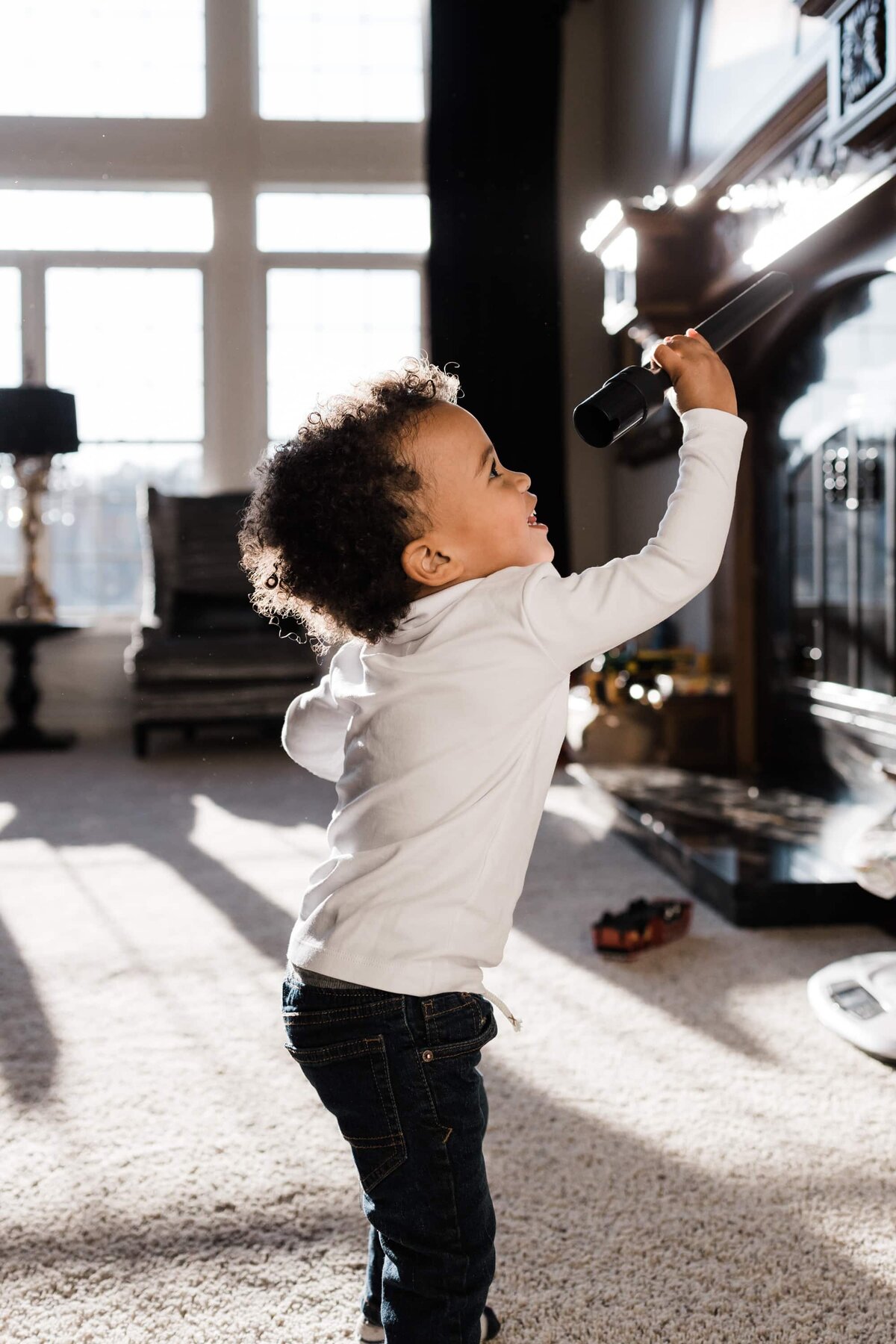 A young child reaching up toward a television remote with sunlight streaming through windows in the background during a DIY family photoshoot.