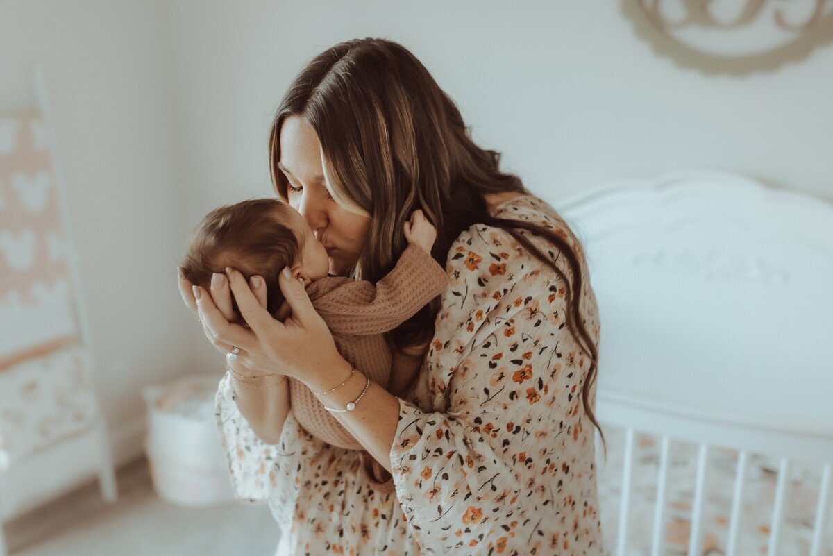 In a cozy nursery, a woman in a floral dress gently kisses a baby on the forehead, capturing a tender moment that any Macon newborn photographer would cherish.