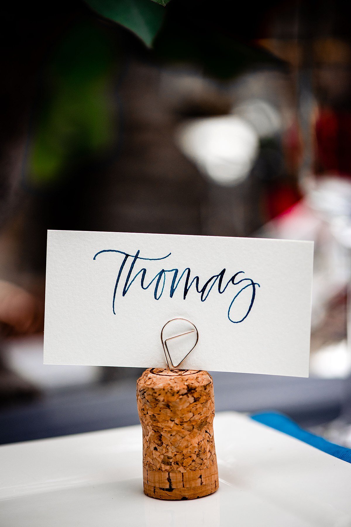 Wine cork seating card stand with a white seating card with teal hand calligraphy sitting on a white square plate with a teal blue napkin.