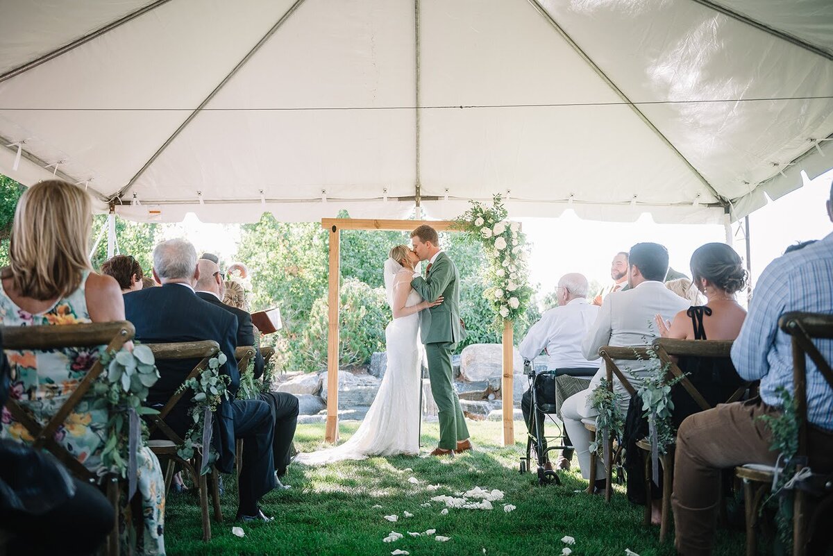 Bride and groom share a kiss during their wedding ceremony in front of a wooden arch with a white and greenery corner arrangement.