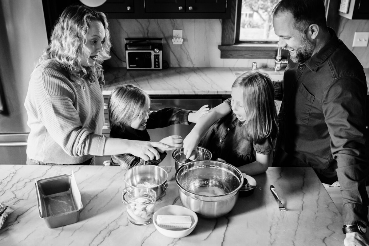 A black and white photo of a family gathered in a kitchen, with the mother and two children actively mixing ingredients in bowls while the father watches with a smile.