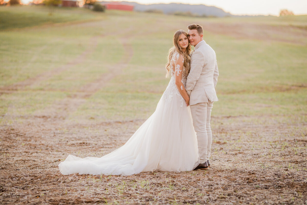 Bride and Groom in open field with train of her dress trailing behind as they lean in cheek to cheek looking at camera