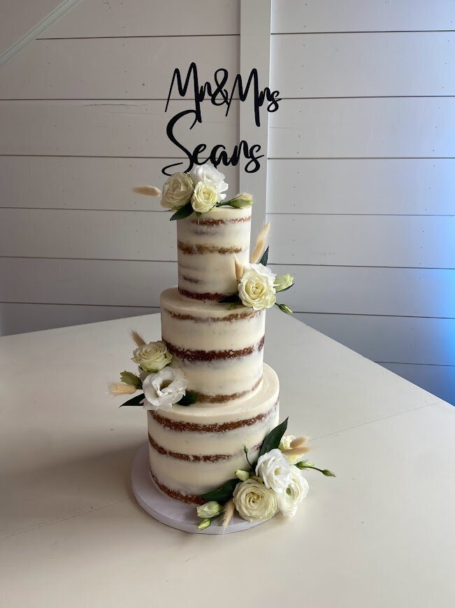 A three tier cake with naked style frosting sits in front of a white paneled wall. Clusters of white and cream fresh flowers alternate sides from top to bottom. There is a black topper that says Mr. & Mrs. Sears in the top tier.