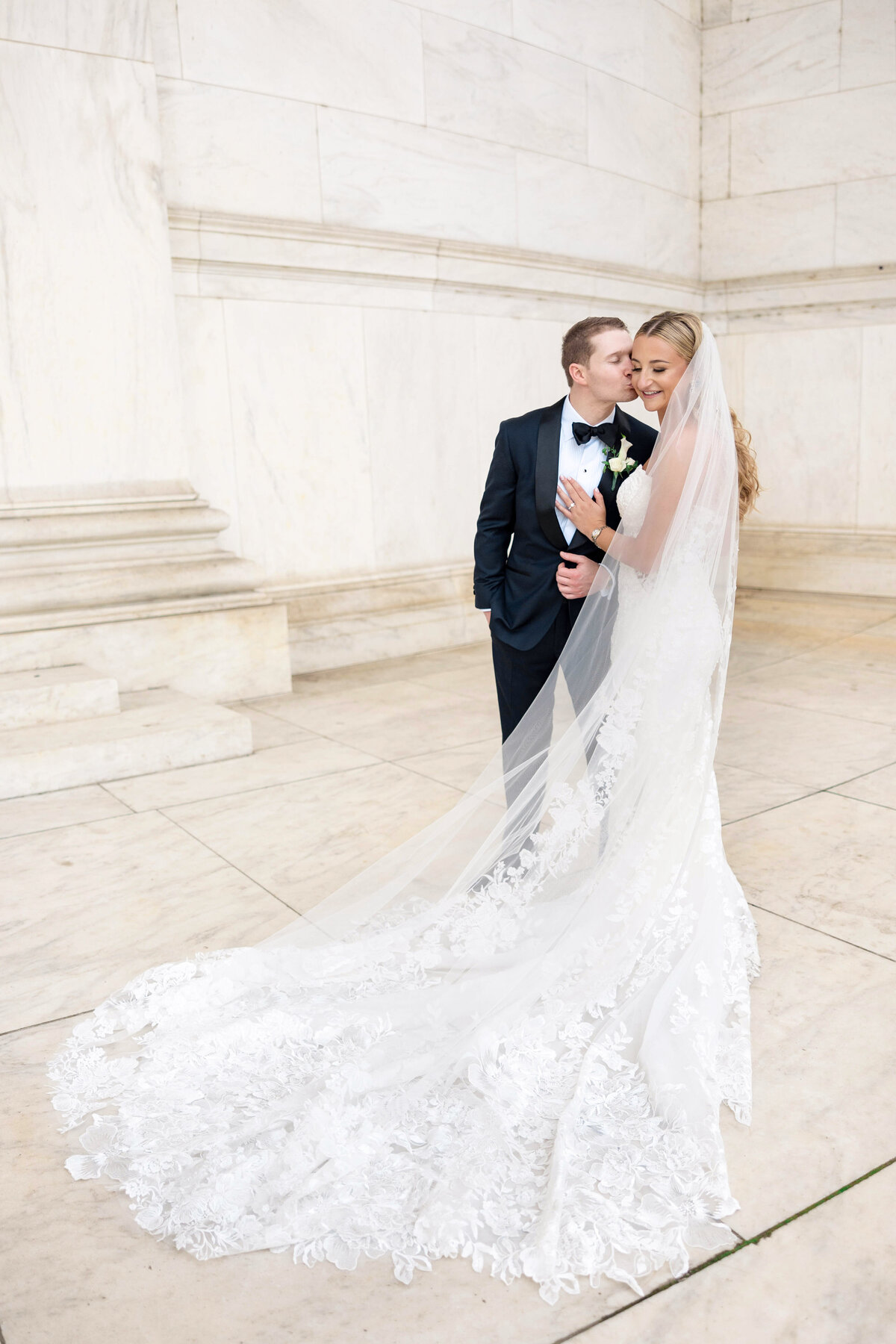 A bride and groom pose affectionately in an elegant setting. The bride wears a long, lace-trimmed wedding gown with a veil, while the groom is in a formal suit with a boutonniere. The background features a marble-like wall.